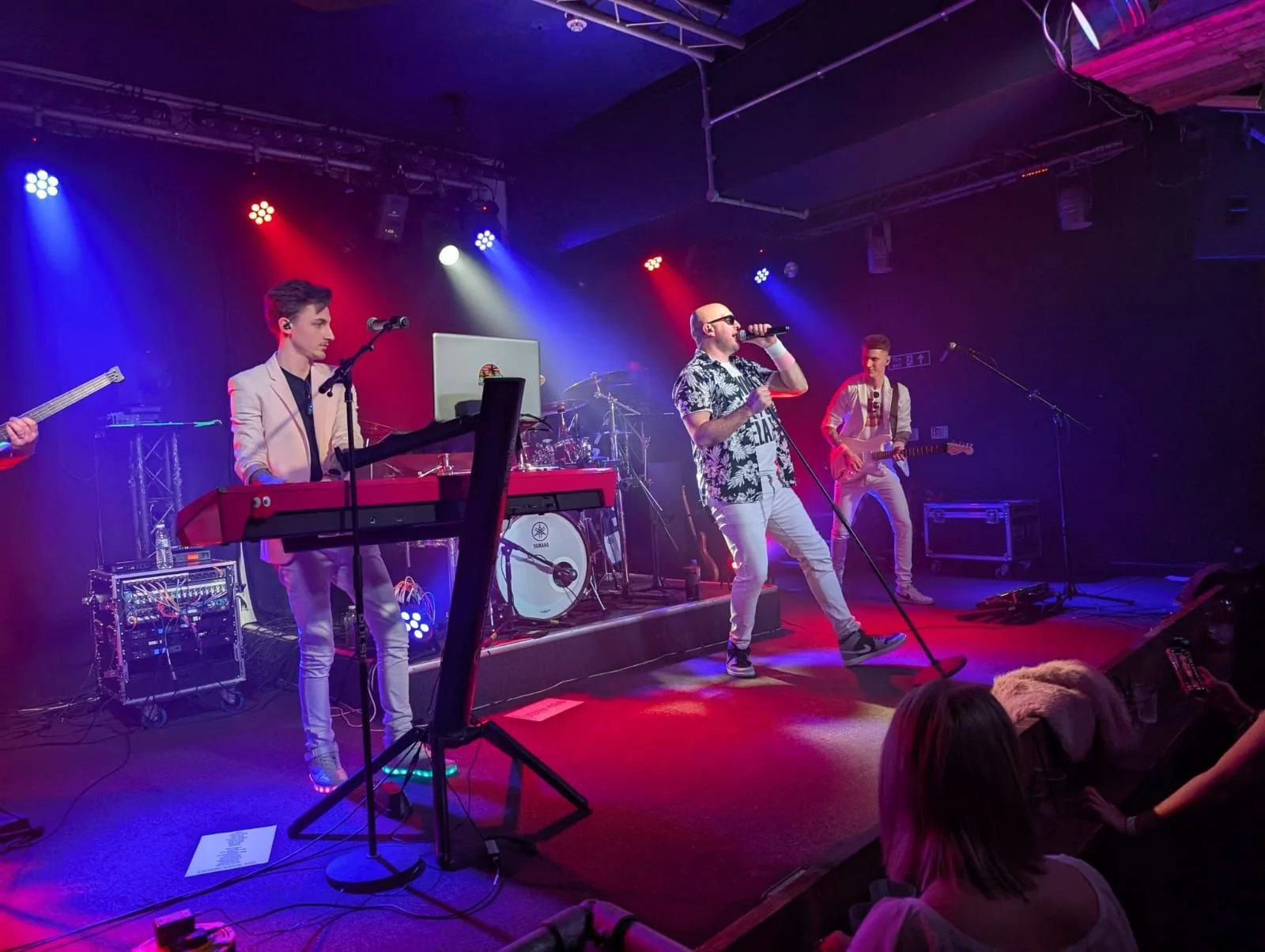 A band performing on stage with a singer, keyboardist, guitarist, and drummer. The stage is lit with colorful lights, and an audience watches the show.