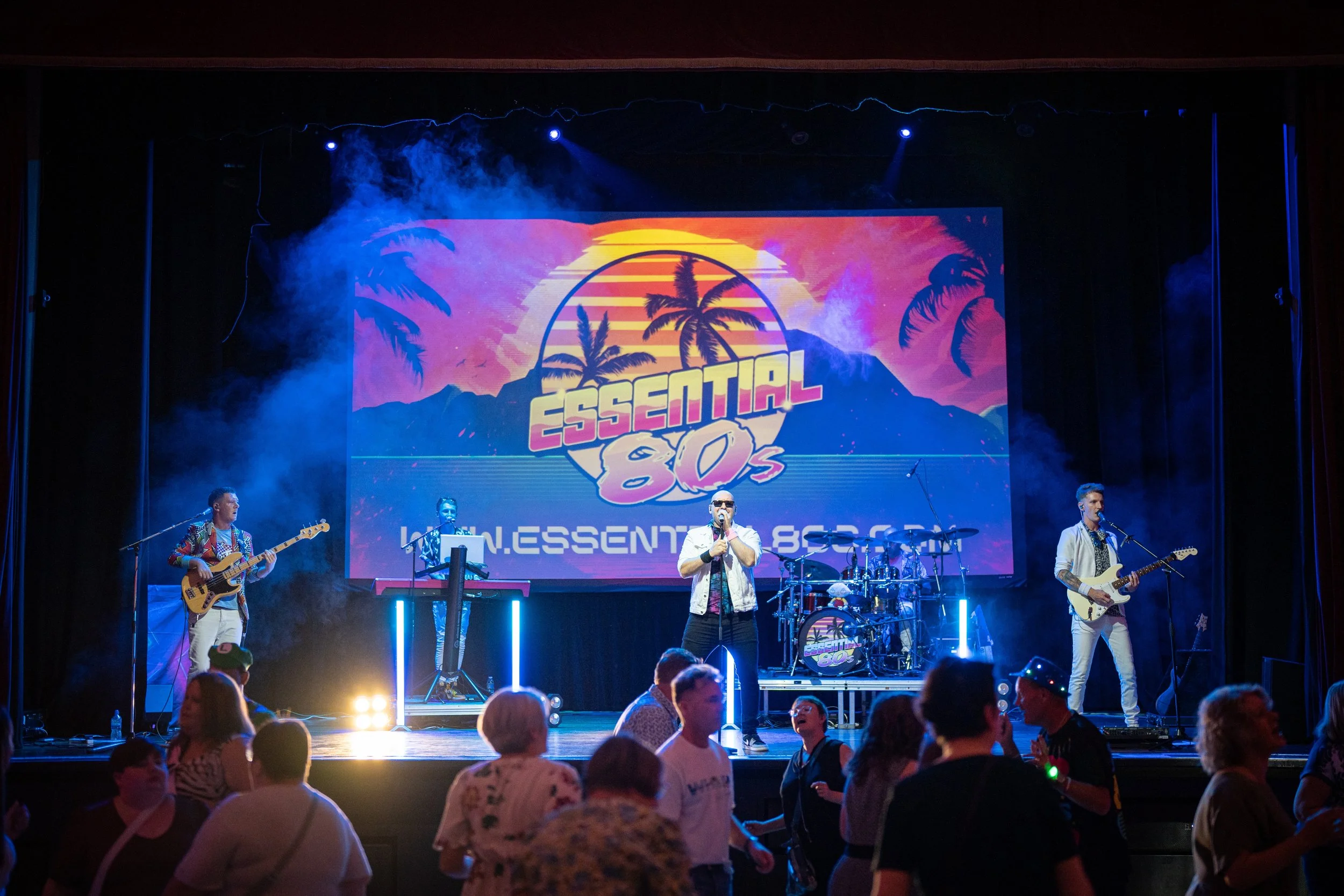 A band performing on stage at the Essential 80s event with a colorful retro sunset backdrop featuring palm trees and mountains, with several people dancing in front of the stage.