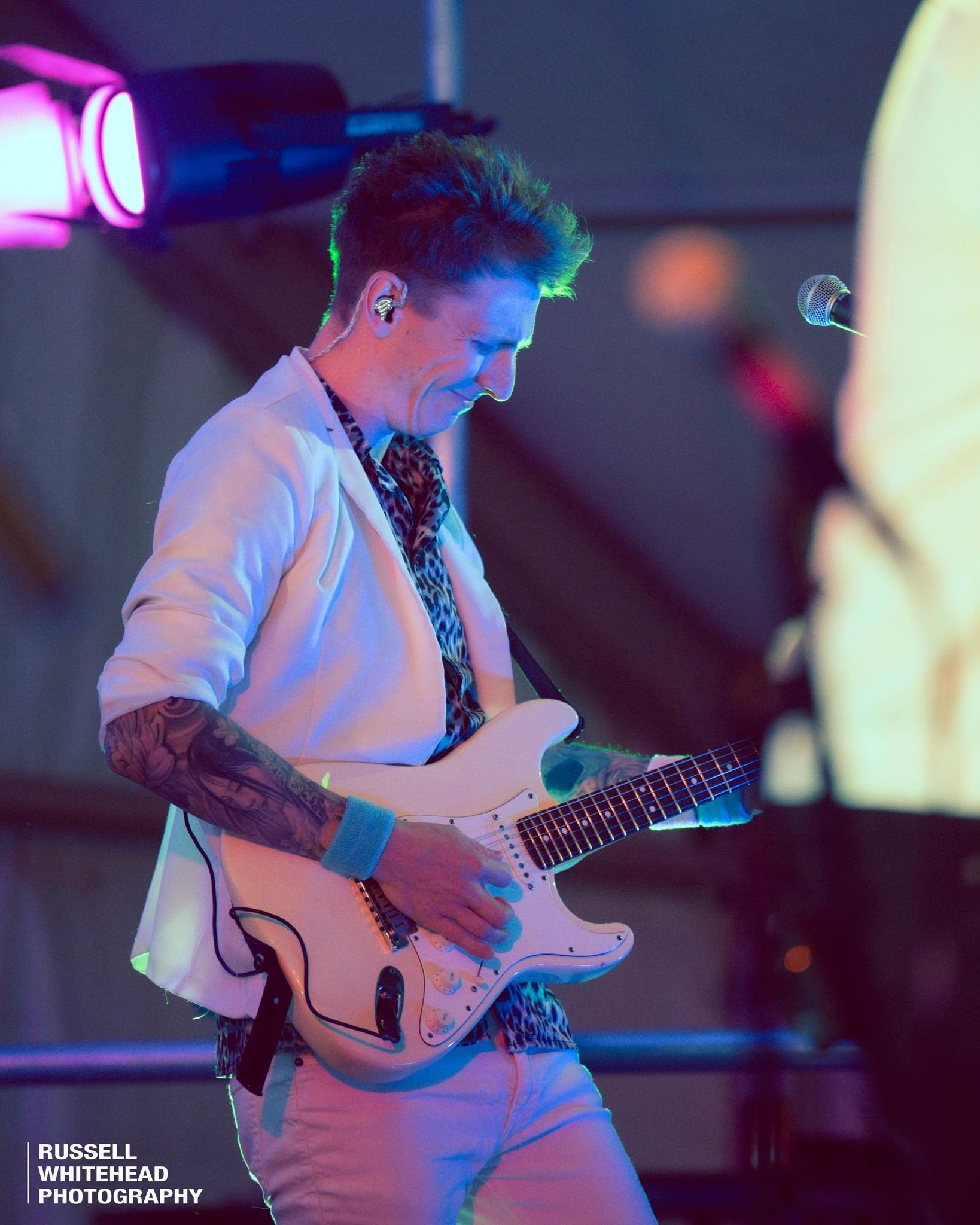 A male guitarist performing on stage, playing an electric guitar. He has dark, styled hair, tattoos on his right arm, and is dressed in a white blazer, leopard print shirt, and white pants. The scene is illuminated with colorful stage lights.
