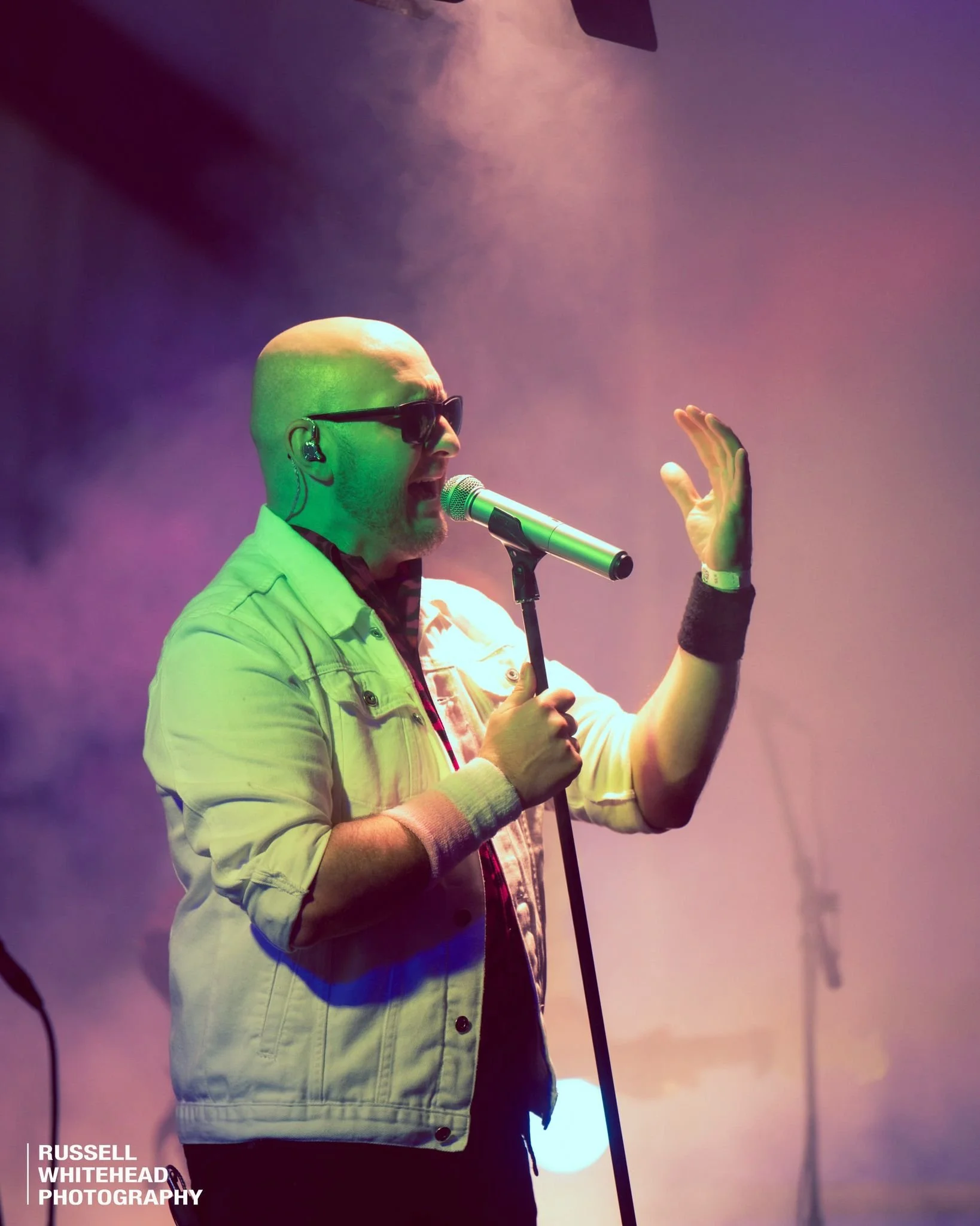 A man with a bald head, sunglasses, and a beard, singing into a microphone on stage. He is wearing a light denim jacket, a black wristband, and a plaid shirt. The stage is filled with colorful purple, pink, and green lights, with fog or smoke effects