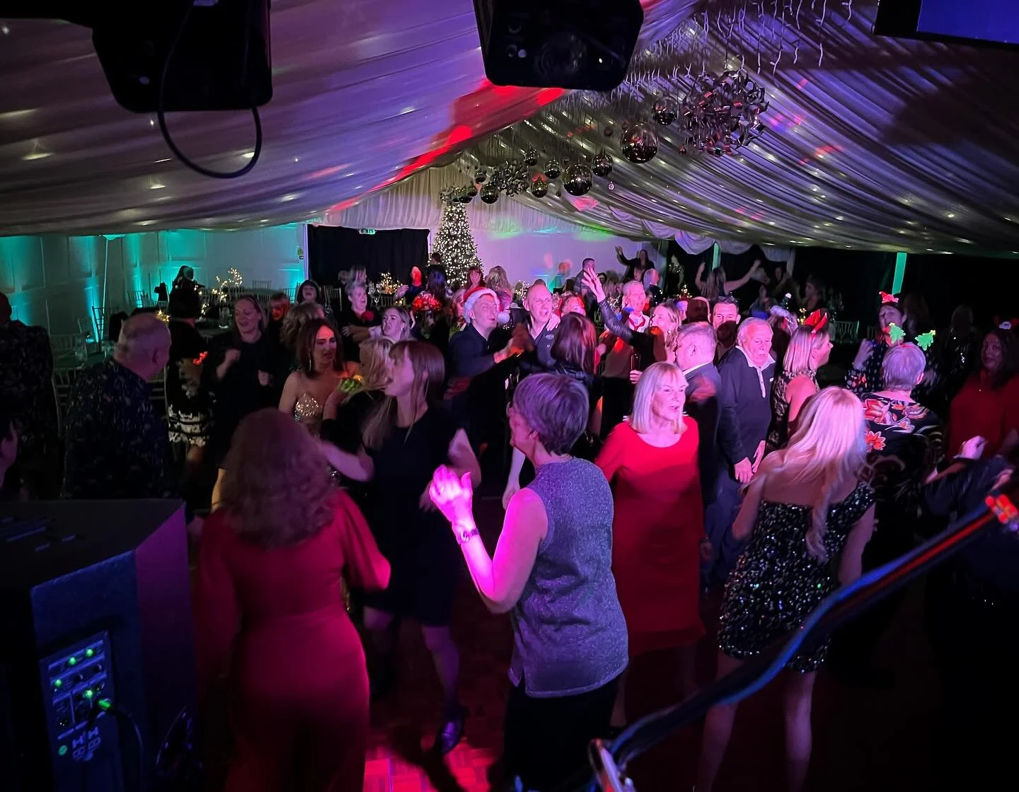 People dancing and celebrating at a holiday party in a decorated venue with Christmas lights and a Christmas tree.