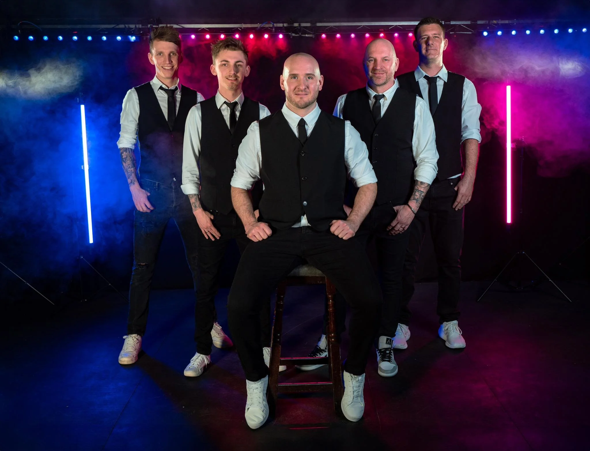 Six men standing together outdoors in front of a concrete wall and a Ferris wheel in the background. They are smiling and dressed in casual summer clothing.