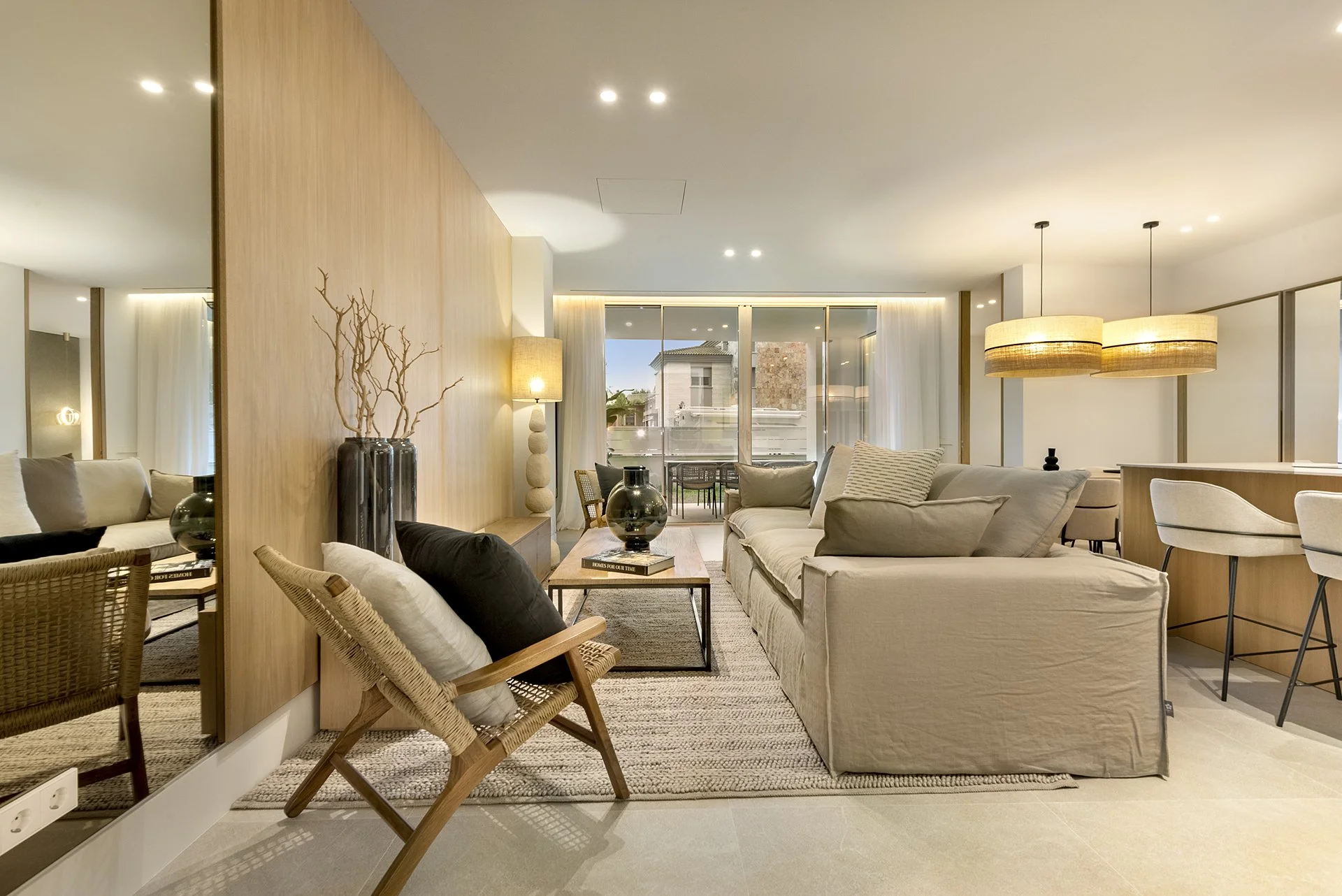 Modern living room with beige sofas, a coffee table with black vases, wooden accents, and large windows with white curtains, decorated with neutral tones and contemporary décor.