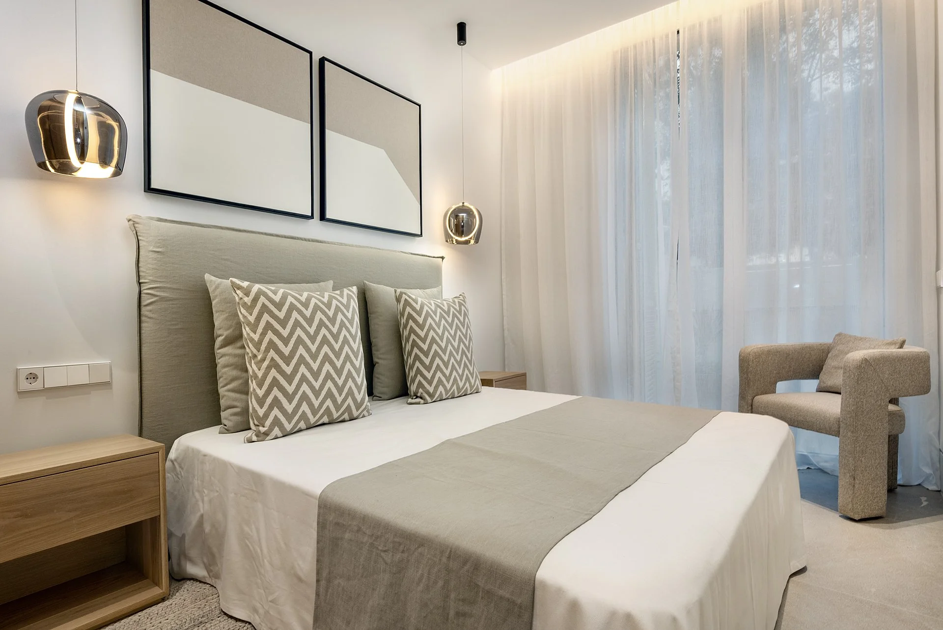 Modern bedroom with a bed, three decorative pillows, two wooden nightstands, hanging pendant lights, abstract wall art, sheer curtains, and a beige armchair near the window.