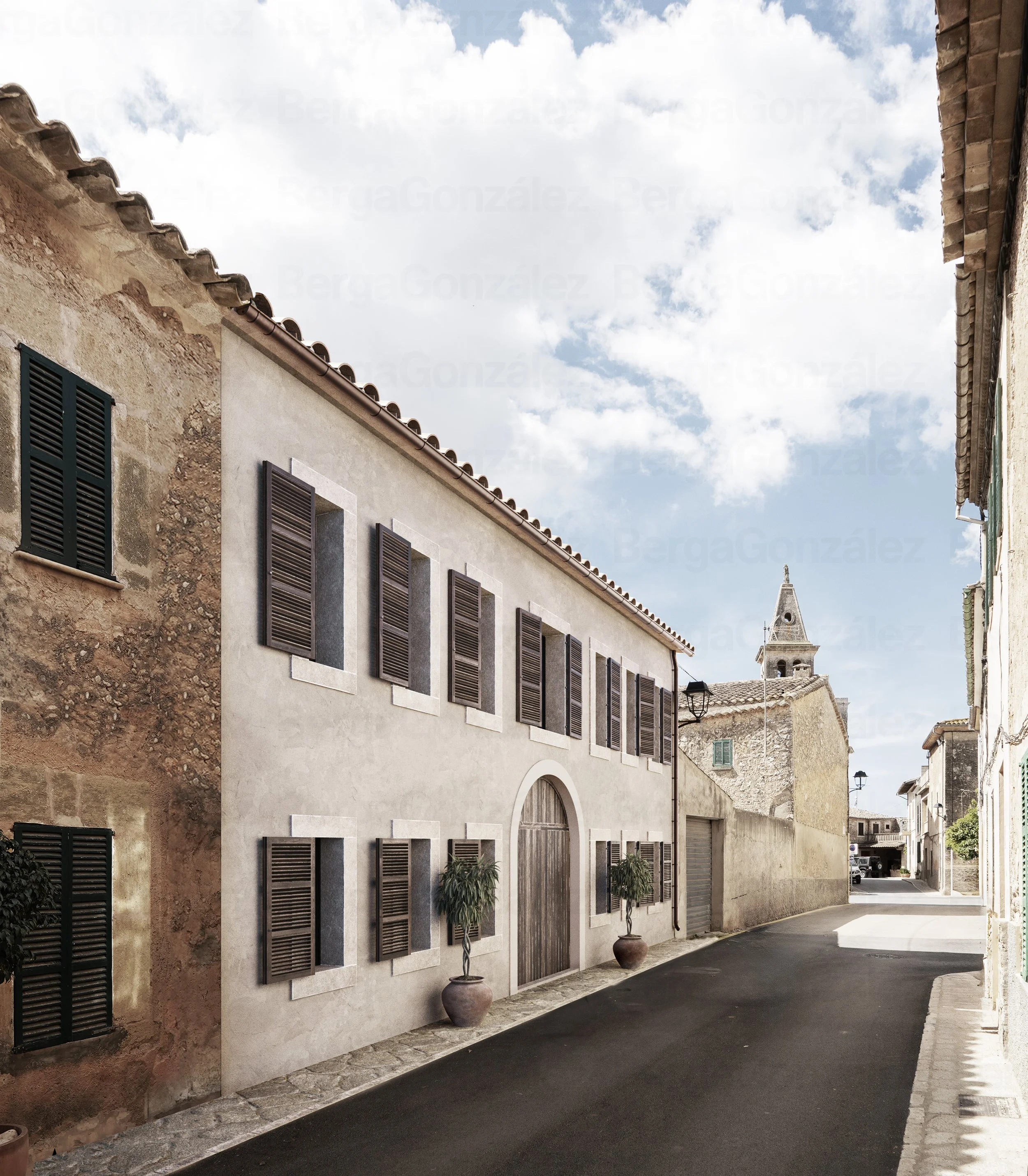 A narrow street in a Mediterranean town with old stone and stucco buildings, some with wooden shutters, and a church steeple in the background under a partly cloudy sky.