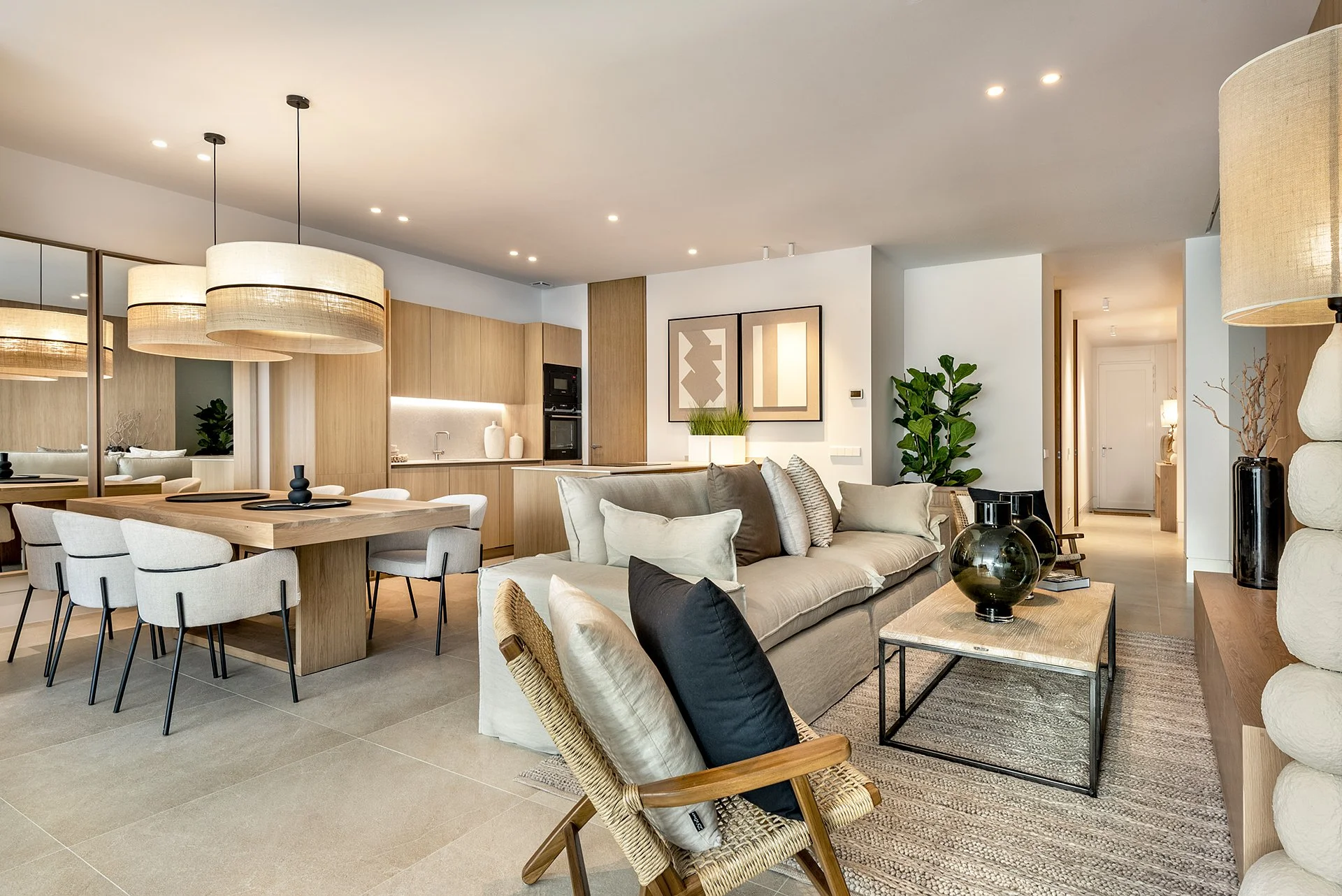 Living room and dining area with beige sofa, armchairs, coffee table, dining table with chairs, wooden kitchen cabinets, pendant lights, artwork on wall, and decorative plants.