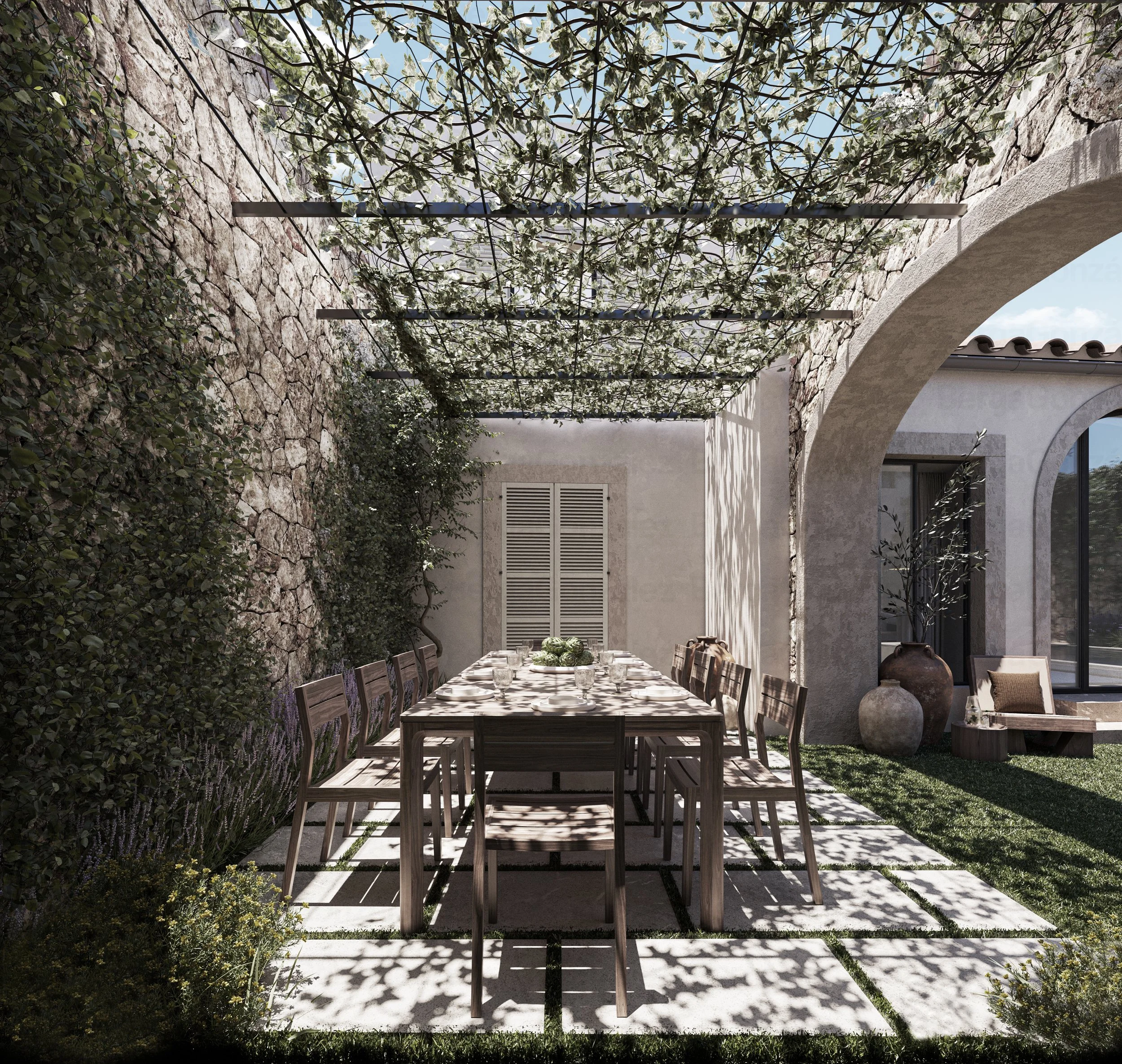 Outdoor dining area with a wooden table and chairs shaded by a vine-covered pergola, stone wall on one side, and a house with arches and potted plants in the background.