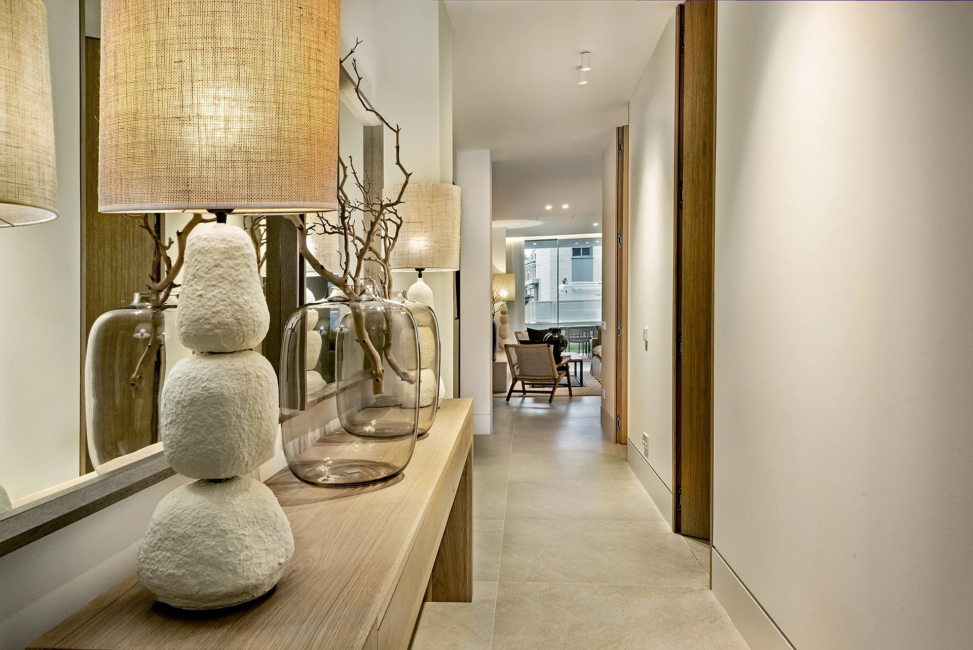 Interior view of a modern hallway with a wooden console table decorated with table lamps, glass vases, and branch arrangements, leading to a living area with seating and large windows.