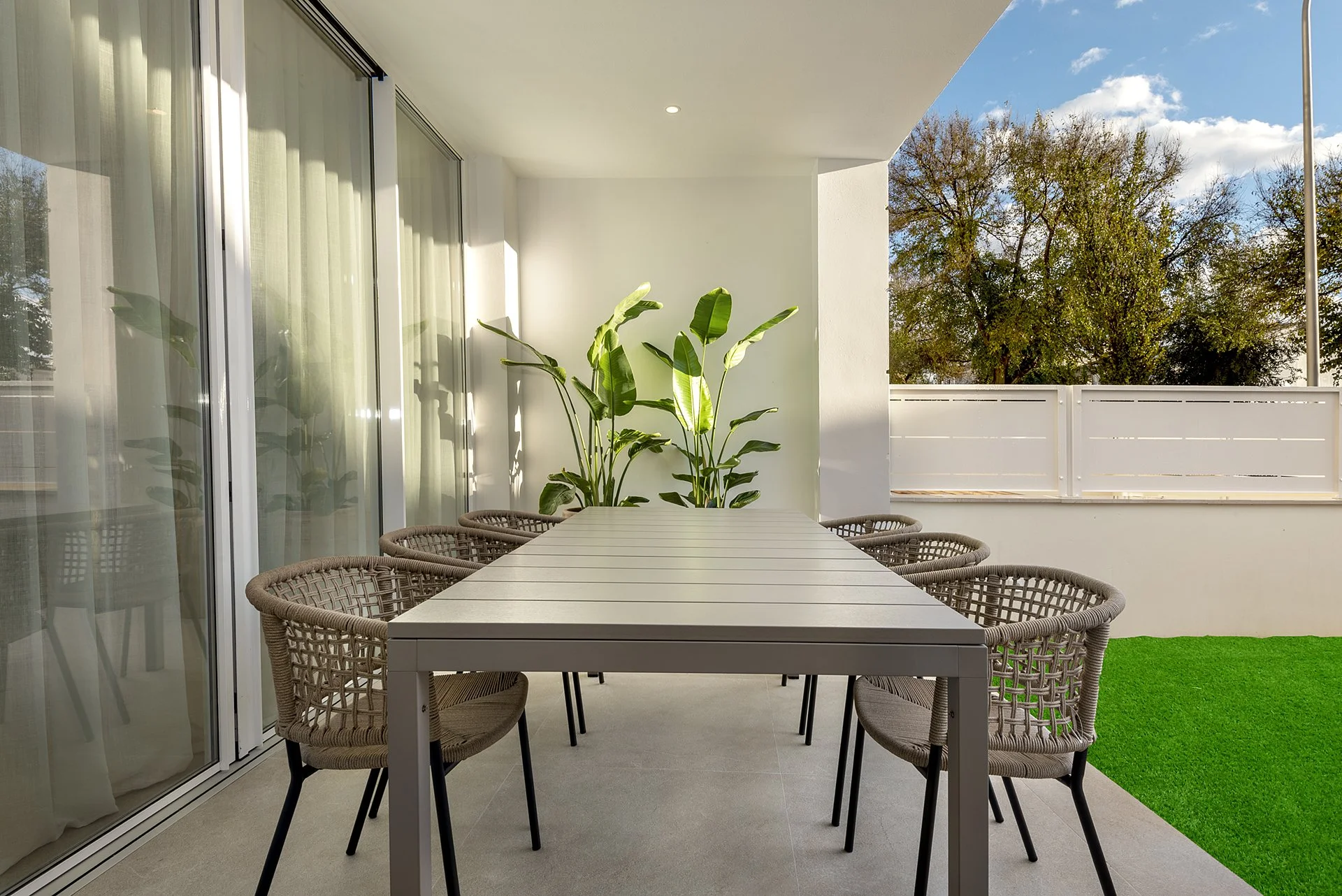 Outdoor balcony with a long white table, eight wicker chairs, potted plants, glass doors, artificial grass patch, and wooden fence, with trees and a cloudy sky in the background.