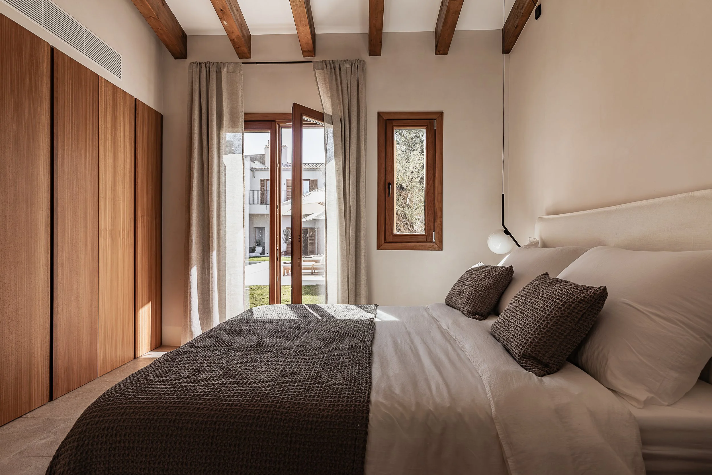 A bedroom with a large bed, wooden ceiling beams, a glass door with curtains leading outside, and two windows with wooden frames.