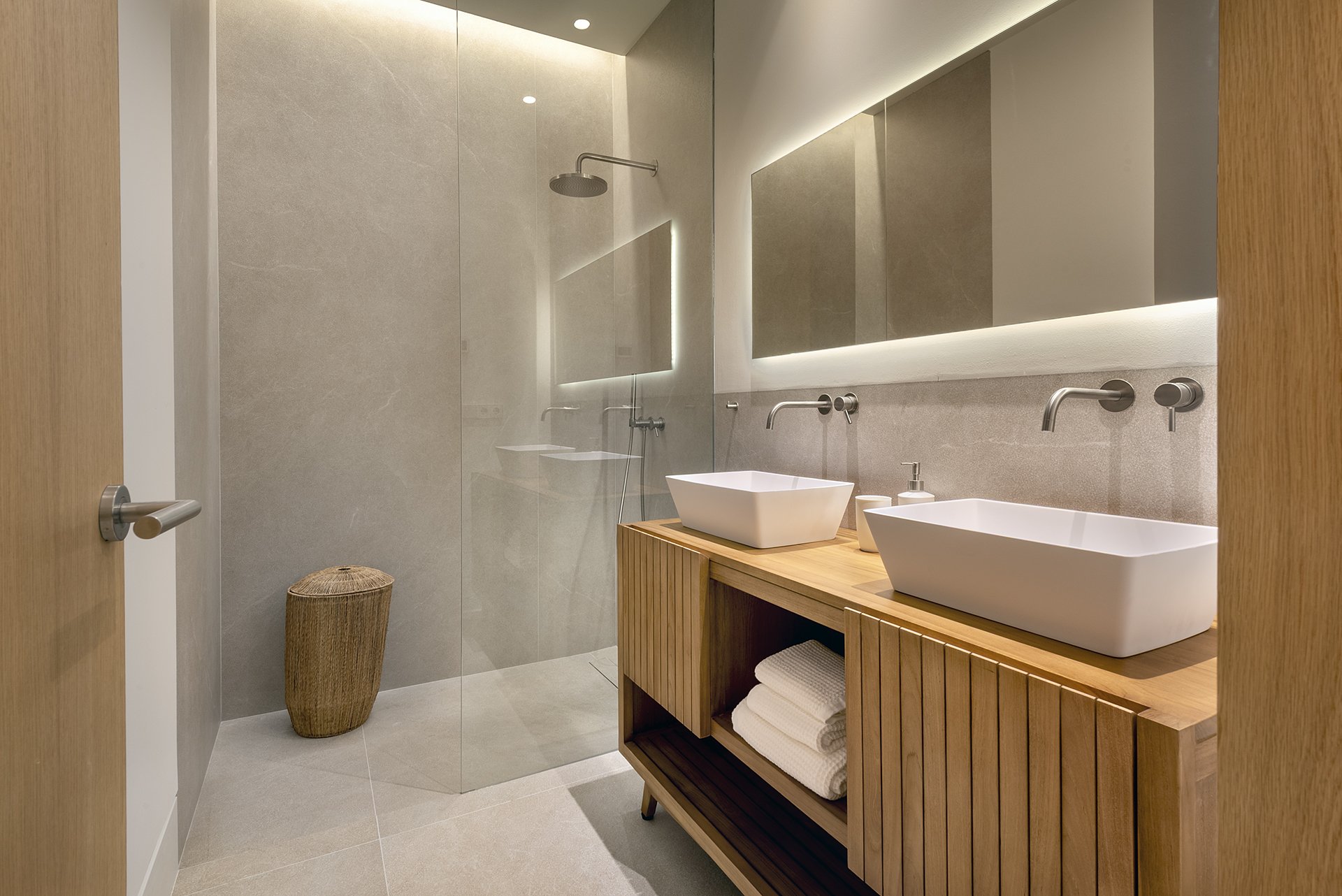 Modern bathroom featuring a walk-in shower with a rain showerhead, two vessel sinks on a wooden vanity, large illuminated mirror, beige tiled walls and floor, and a woven laundry basket.