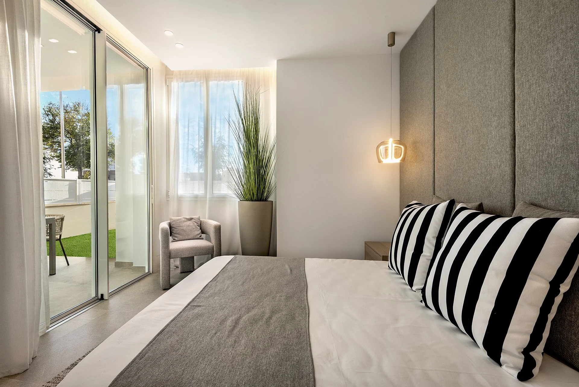 Modern bedroom with a bed, gray headboard, black and white striped pillows, a hanging light fixture, large window with sheer curtains, a tall plant in a pot, a gray armchair, and a balcony with outdoor furniture.