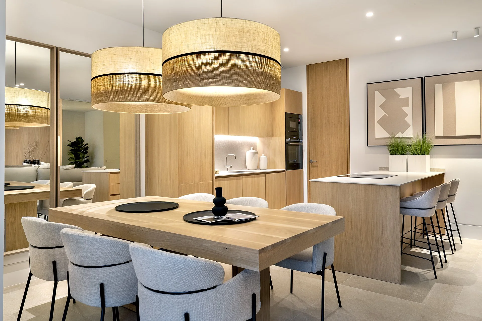 Modern kitchen and dining area with wooden accents, pendant lights, a large dining table with six upholstered chairs, a kitchen island with seating, and artwork with plants above.