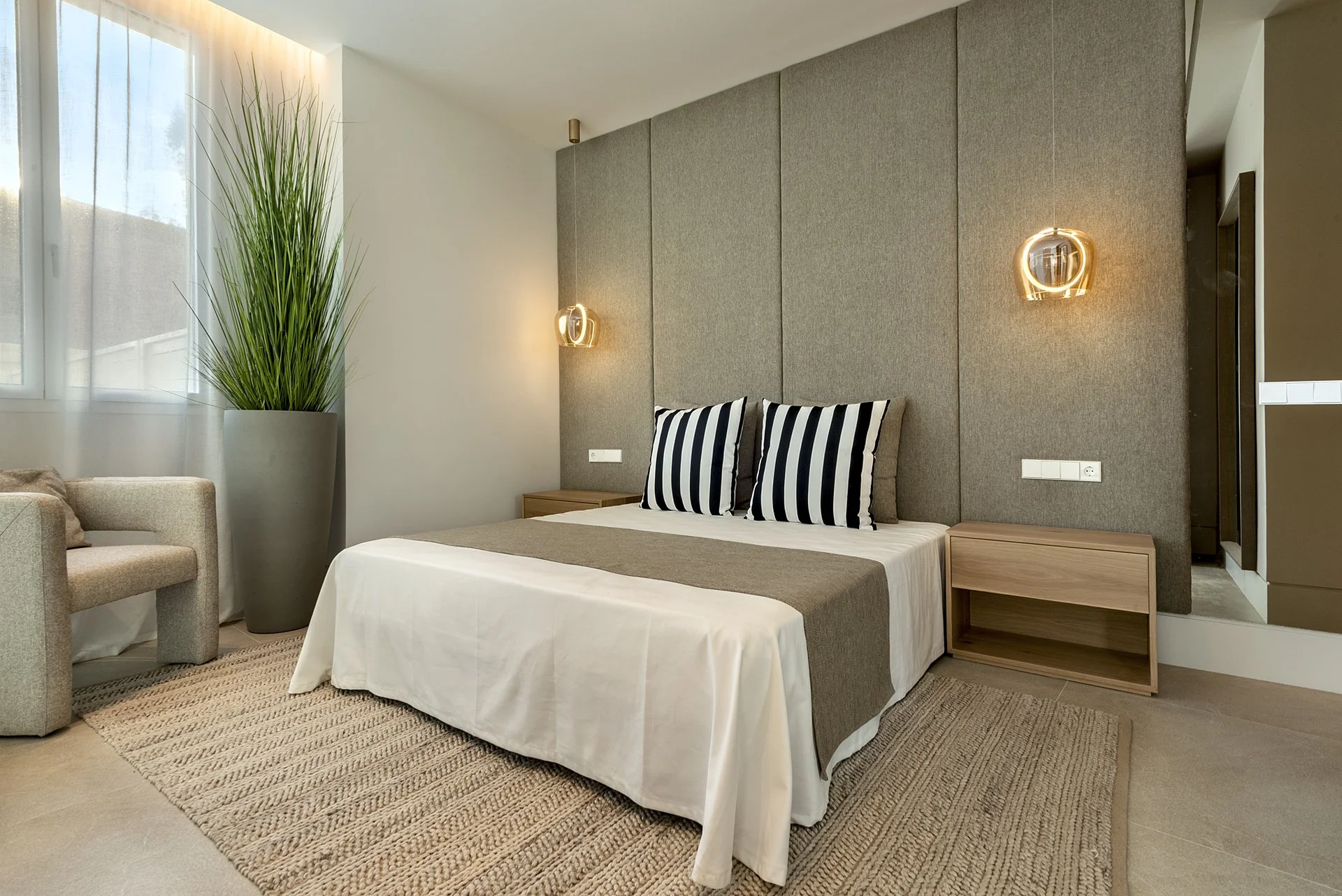 Modern bedroom with a bed featuring striped pillows, a wooden bedside table, a tall green plant in a large gray pot, and wall-mounted pendant lights.