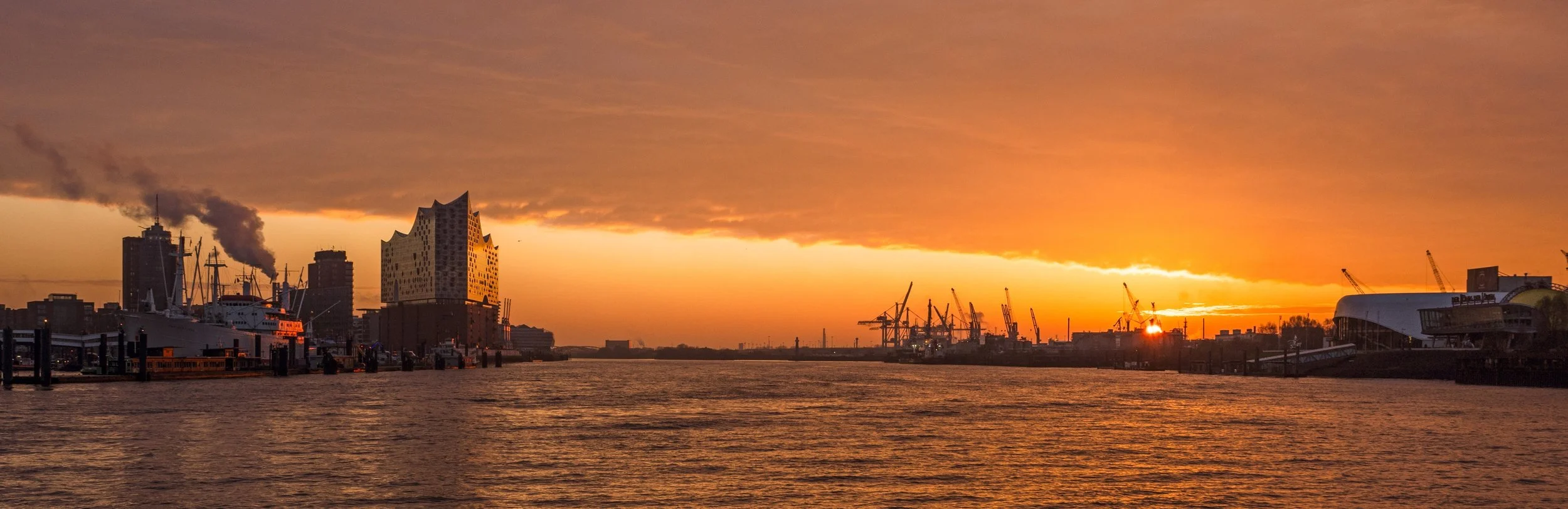 Sonnenuntergang über einem Hafen mit Schiffen, Gebäuden und Krane in Hamburg, Deutschland.
