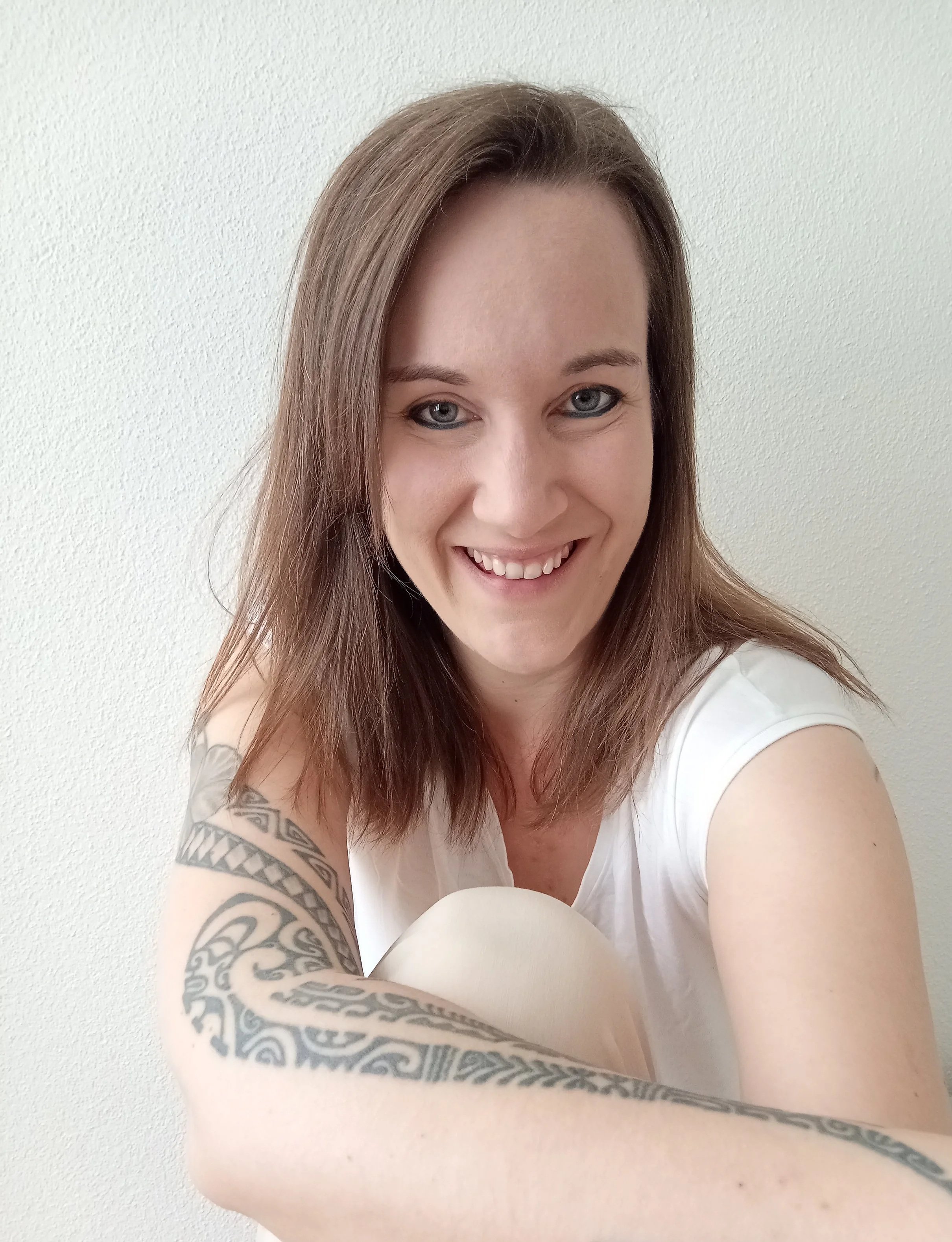 Foto einer lächelnden jungen Frau mit braunen Haaren und Tattoos auf dem Arm, vor einer weißen Wand