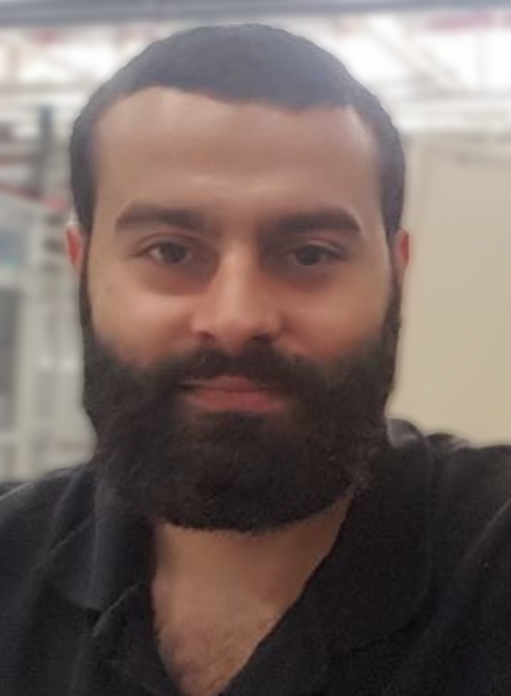 Close-up of a man with short dark hair and a full beard, wearing a black shirt, in an indoor setting.