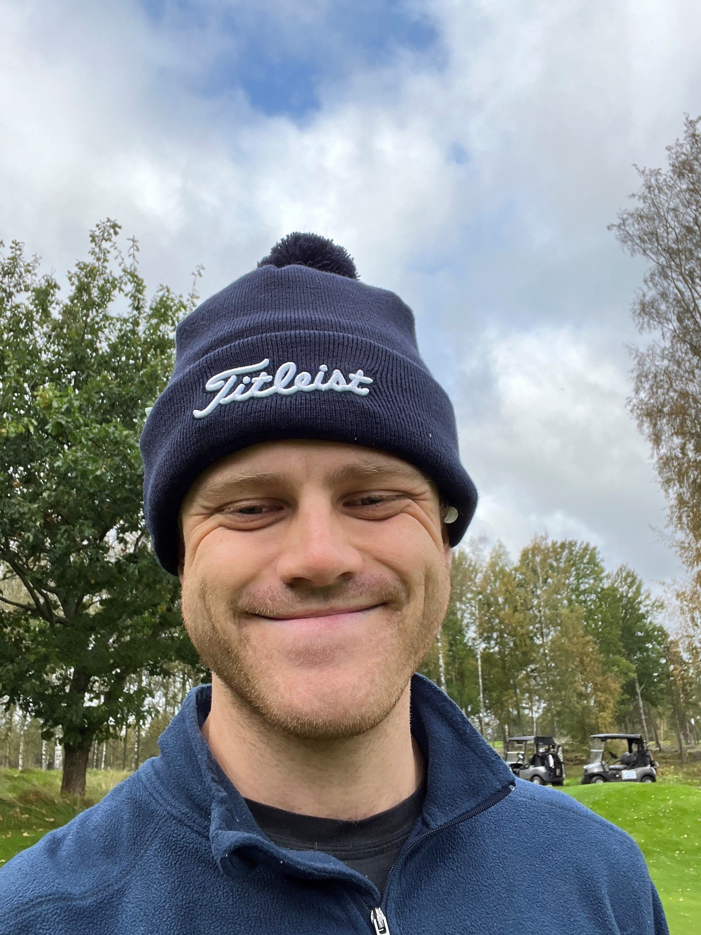 A man smiling outdoors, wearing a navy blue beanie with 'Titleist' embroidered on it, a navy zip-up jacket, with trees and golf carts in the background, under a cloudy sky.