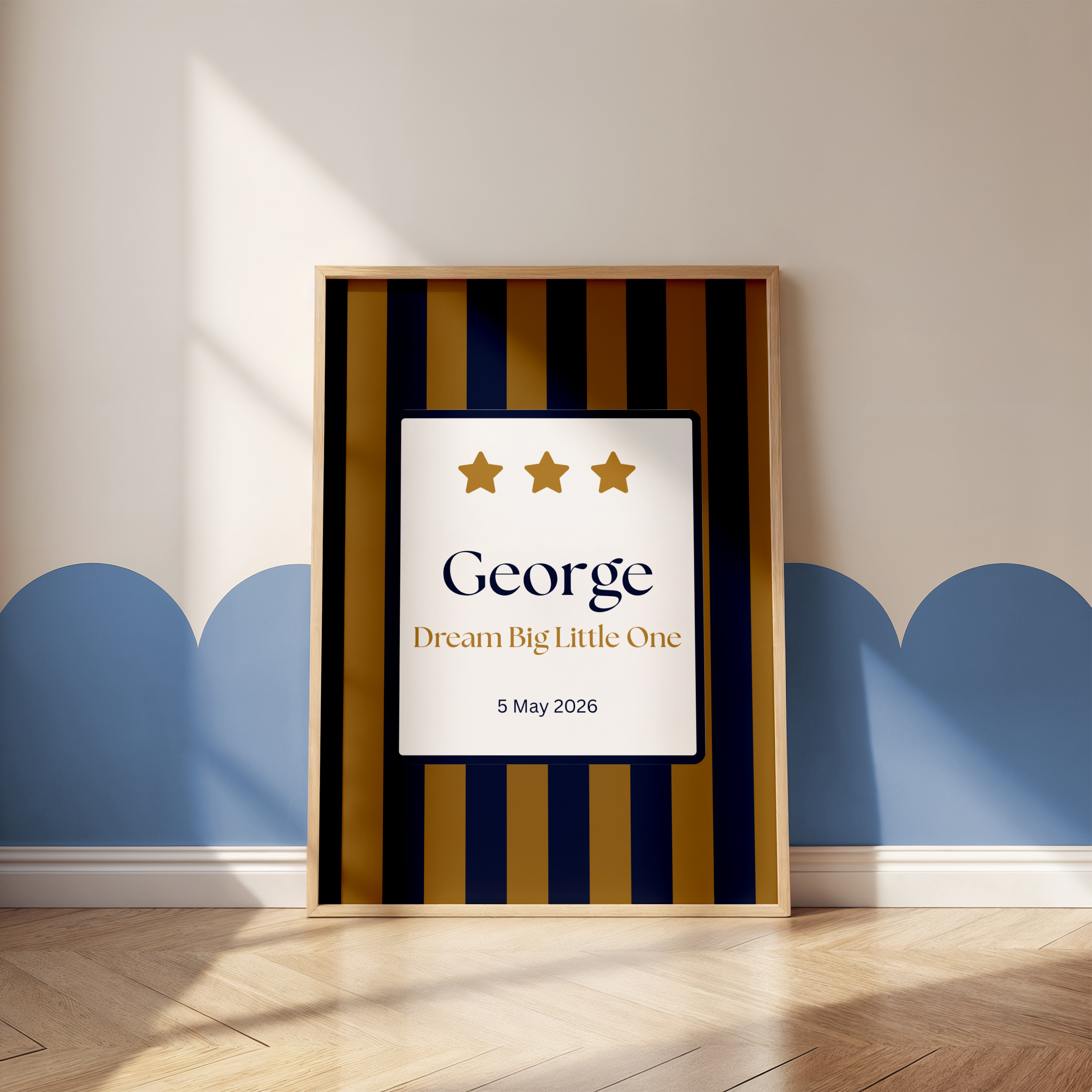 Framed modern artwork with a white background and three gold stars, with the text 'George' and 'Dream Big Little O', dated 5 May 2026, leaning against a wall with a wooden floor.
