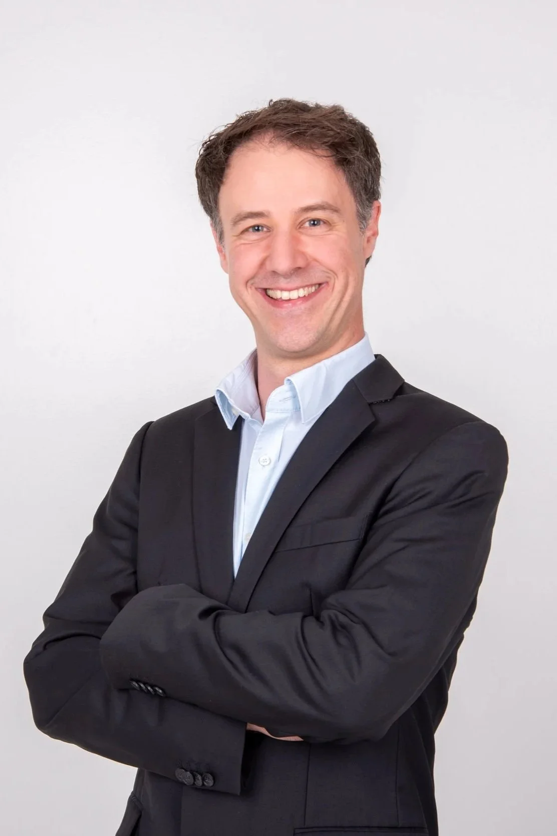 Ein lachender Mann in einem schwarzen Anzug mit gehobener Haltung vor weißem Hintergrund.