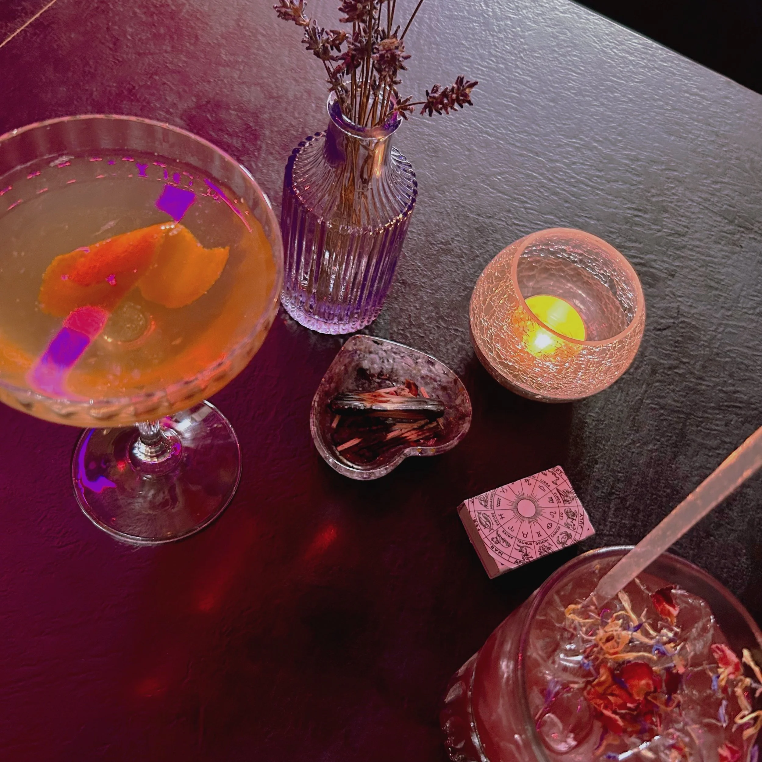 A close-up of a colorful drink with a orange slice garnish in a glass on a dark surface. In the background, there is a purple vase with dried lavender flowers and a lit candle in a textured pink holder, creating a cozy ambiance.
