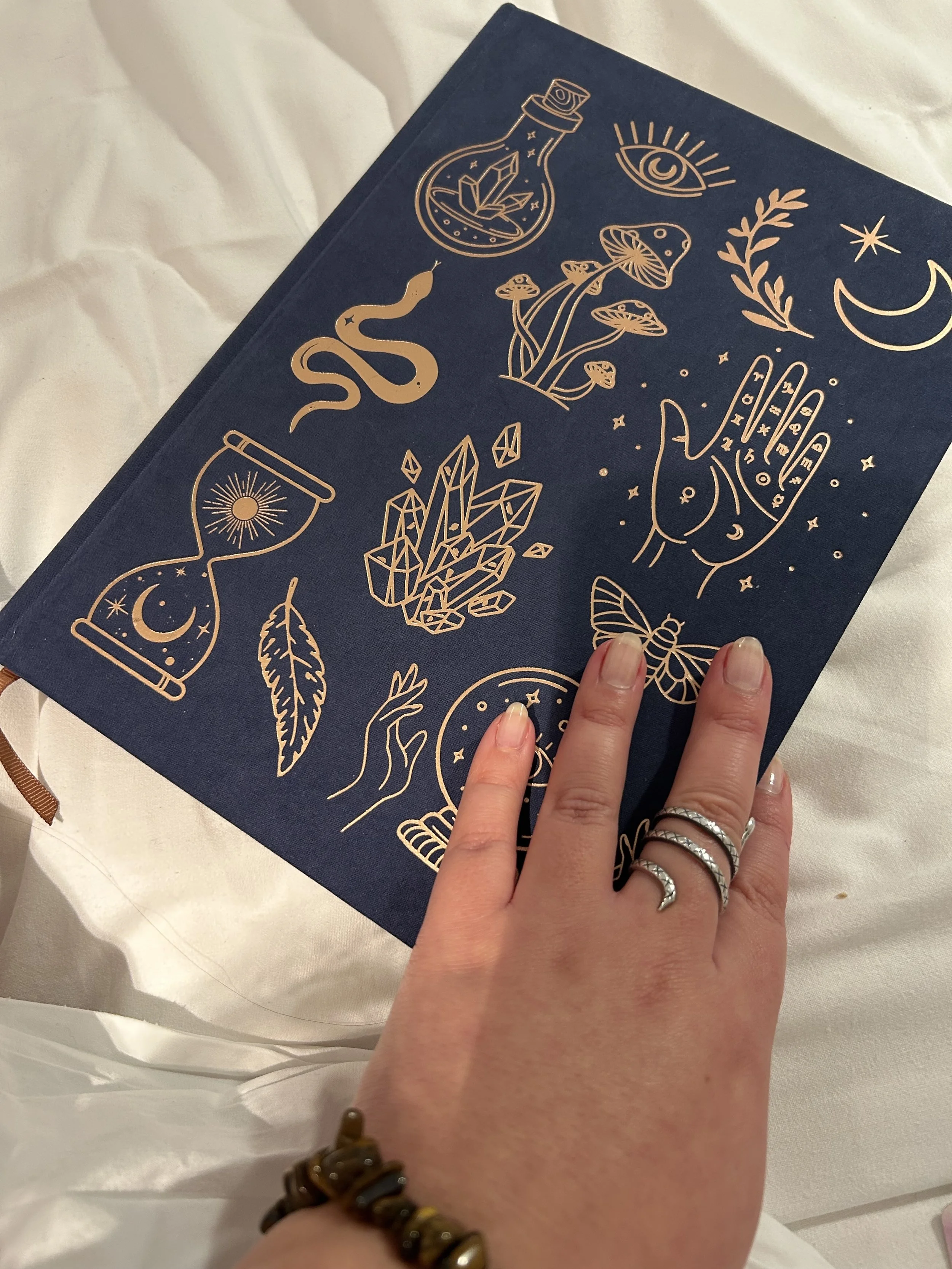Person's hand with rings touching a navy blue notebook with gold mystical line drawings, including an eye, crescent moon, crystals, mushrooms, snake, hourglass, and stars.