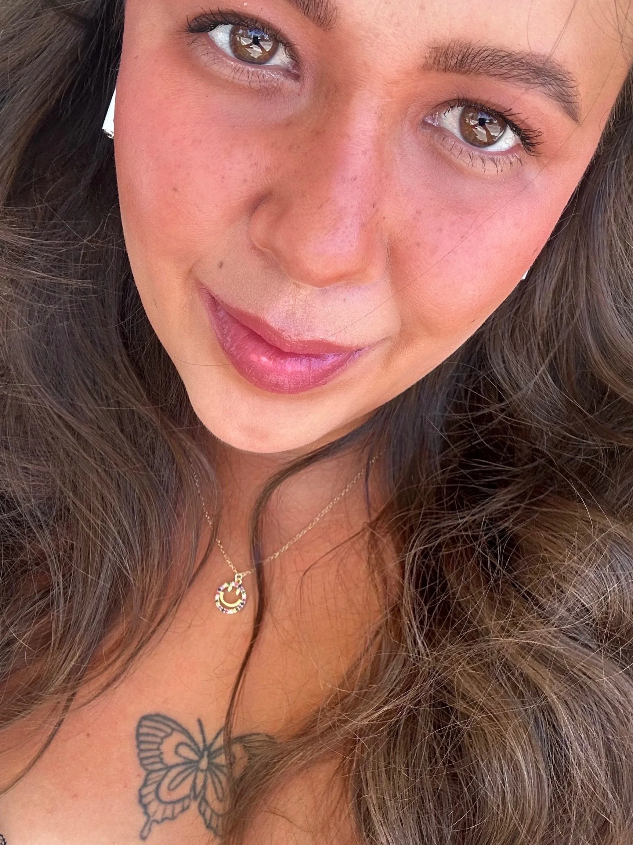 Close-up of a woman's face showing her brown eyes, eyebrows, and a butterfly tattoo on her shoulder with long, wavy brown hair. She is wearing makeup and jewelry including a necklace and earrings.