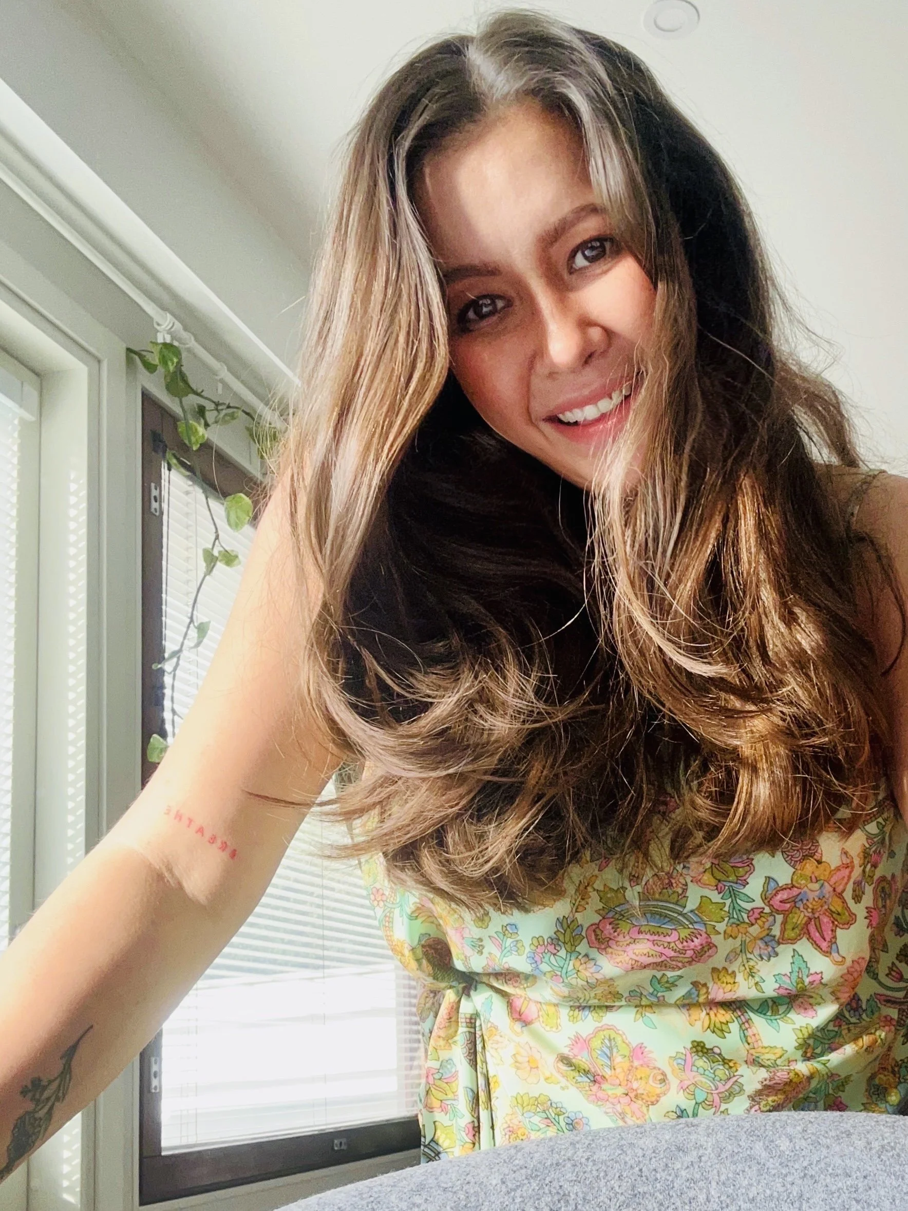 A young woman with long, wavy brown hair smiling at the camera. She has a tattoo on her left arm and a red tattoo on her right wrist. She is wearing a floral dress with bright colors. The background shows a window with white blinds and a climbing plant.