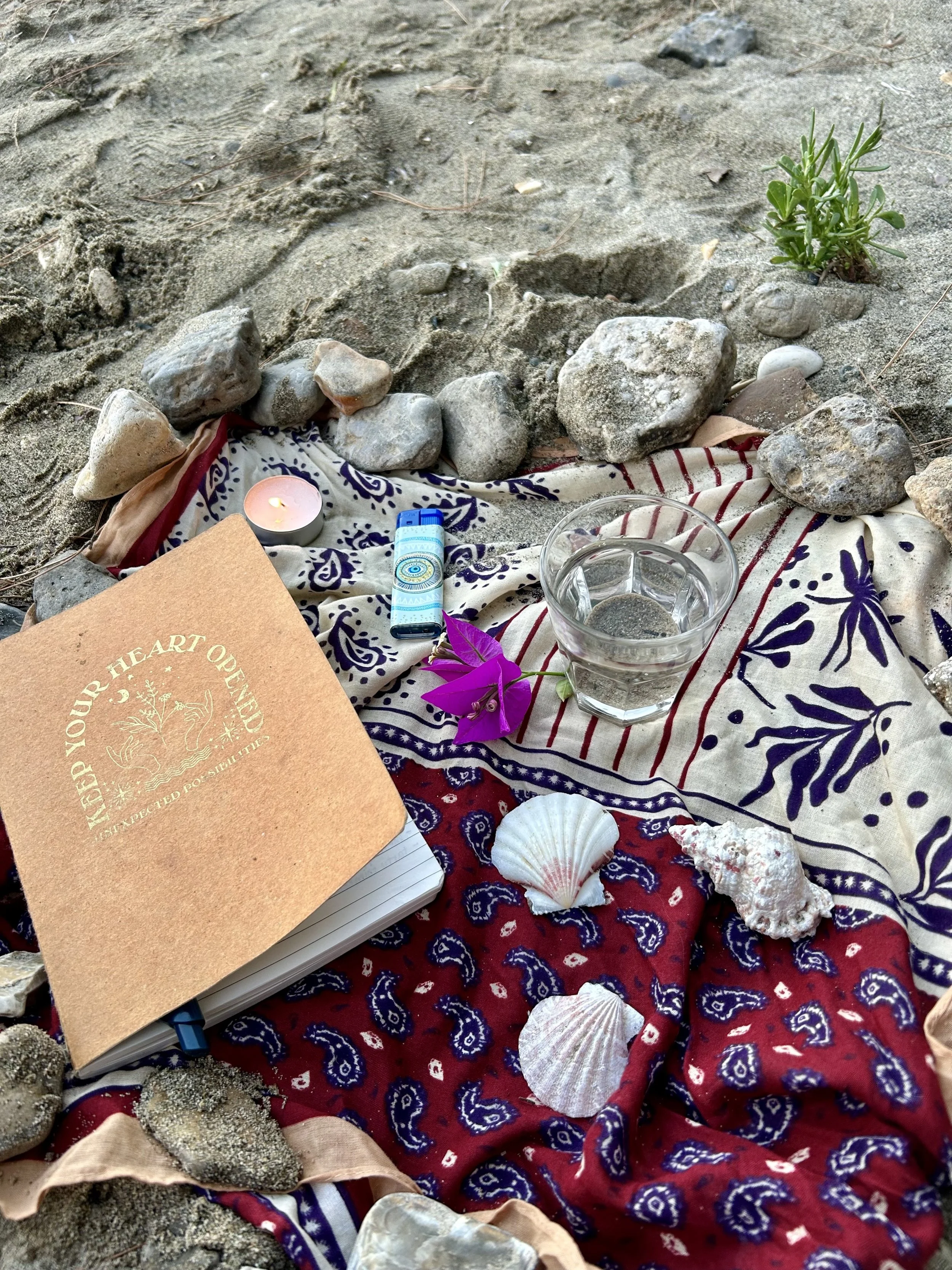 A small personal ritual arranged on a beach: candle, notebook, flowers, and fabric.