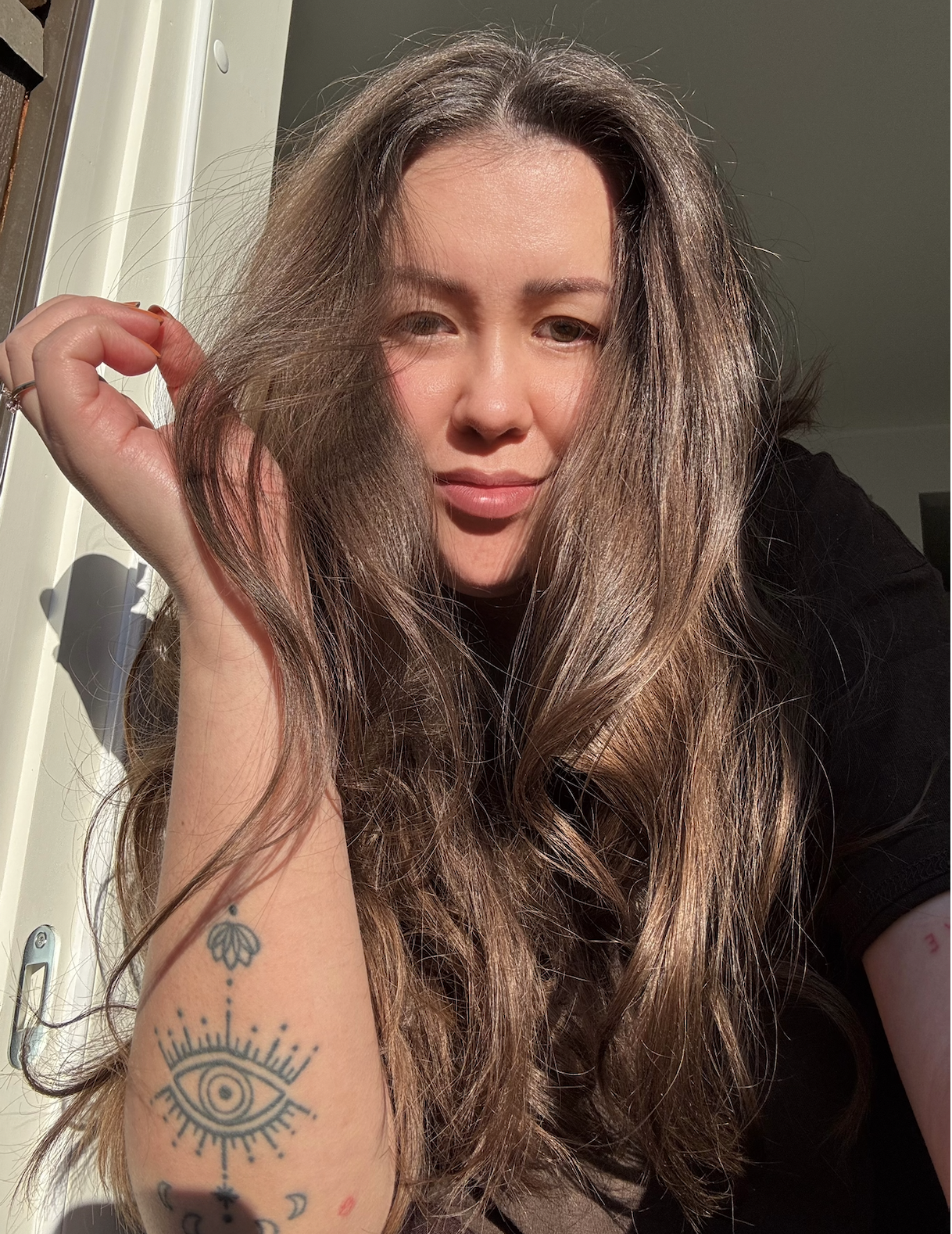 A woman with long, wavy hair and tattoos on her arm, holding her hand near her face, in a room with sunlight streaming in.