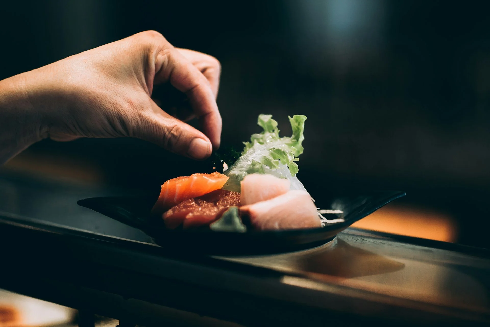 Una mano colocando delicadamente una hoja de lechuga en un plato con sashimi variado sobre una mesa.