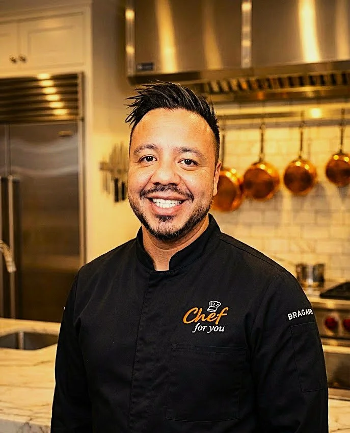 Chef sonriendo en una cocina con utensilios de cobre en la pared y electrodomésticos de acero inoxidable en el fondo.