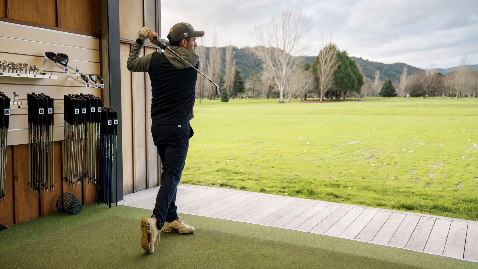 Man practicing golf swing at driving range with golf clubs on wall and open field with trees in background