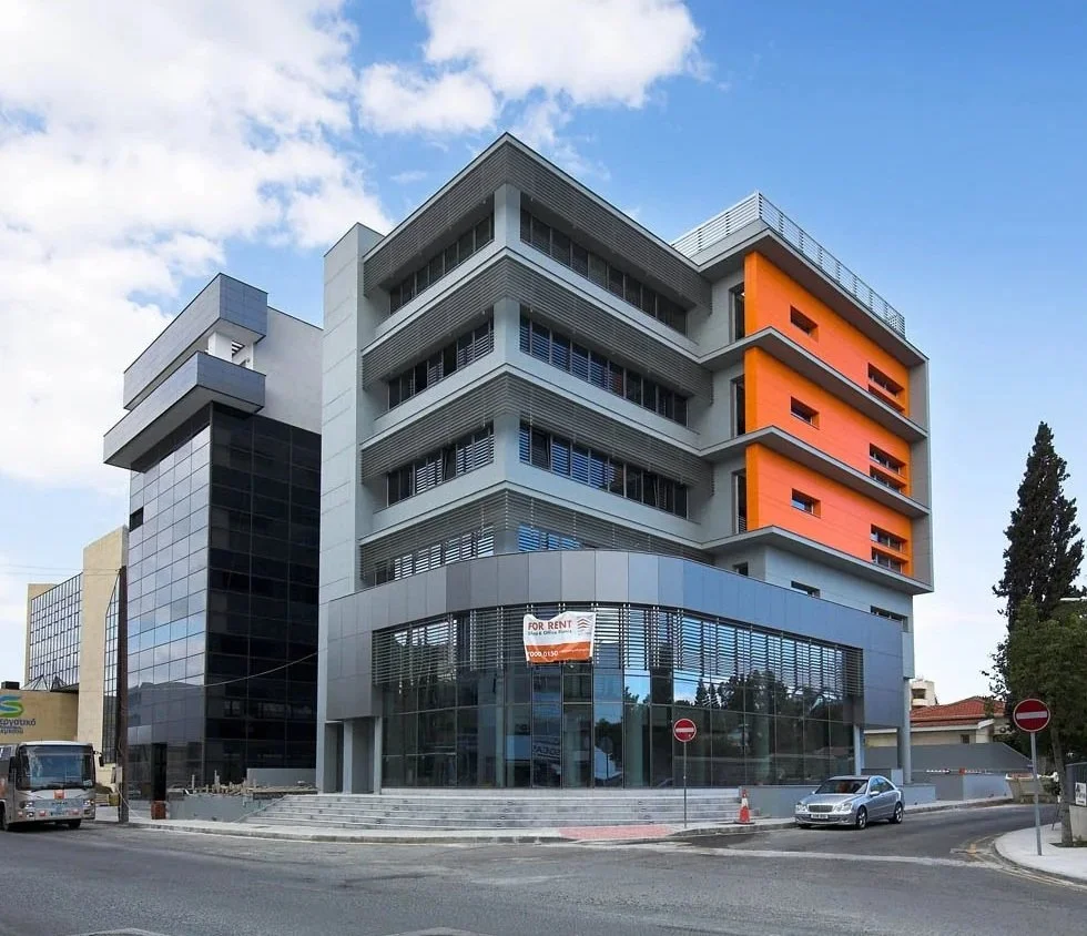 Modern multi-story commercial building with glass windows, orange accent panels, and a "For Rent" sign in front.