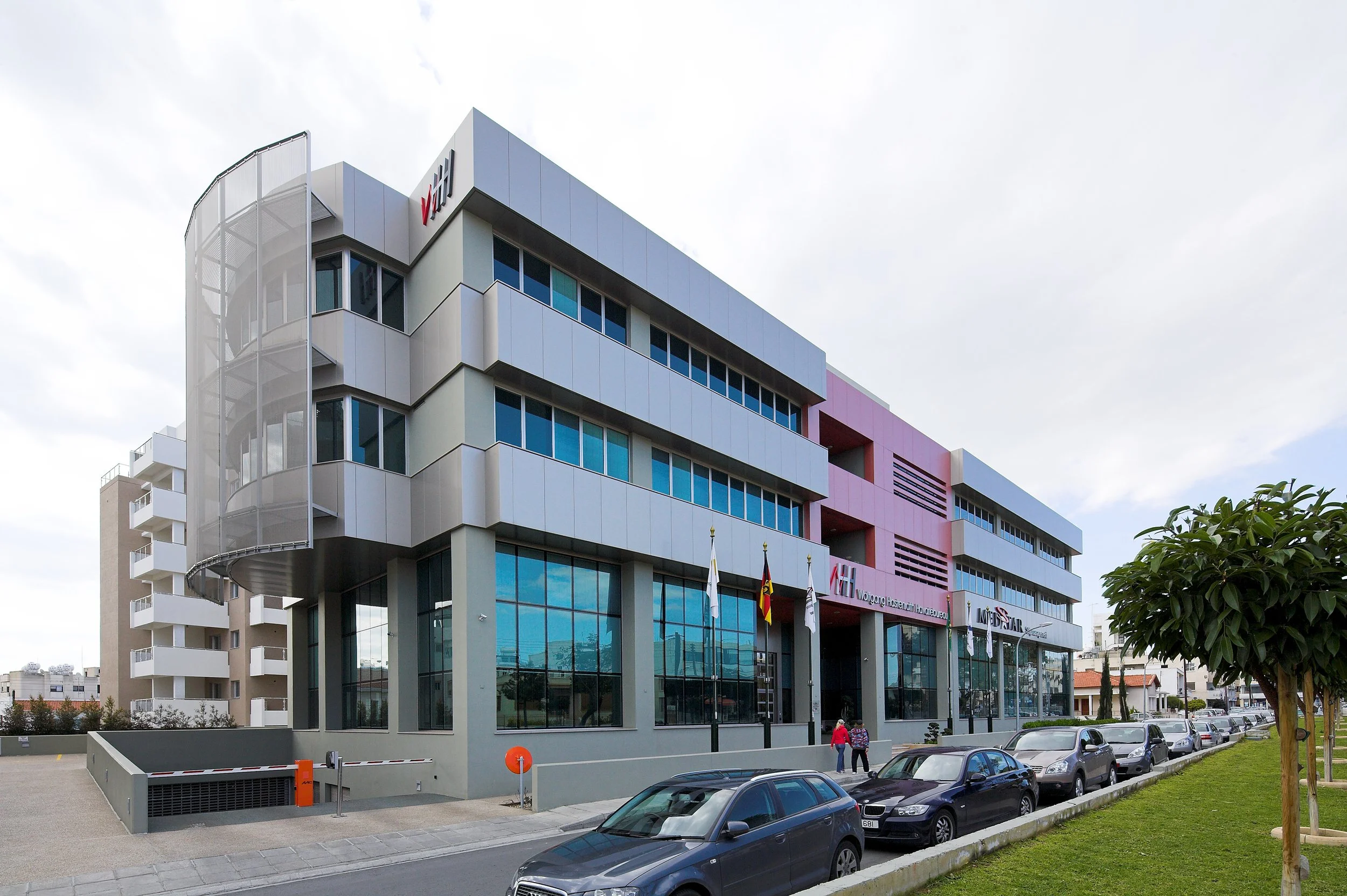 Modern multi-story office building with glass exterior, flags out front, parking lot, and cloudy sky.