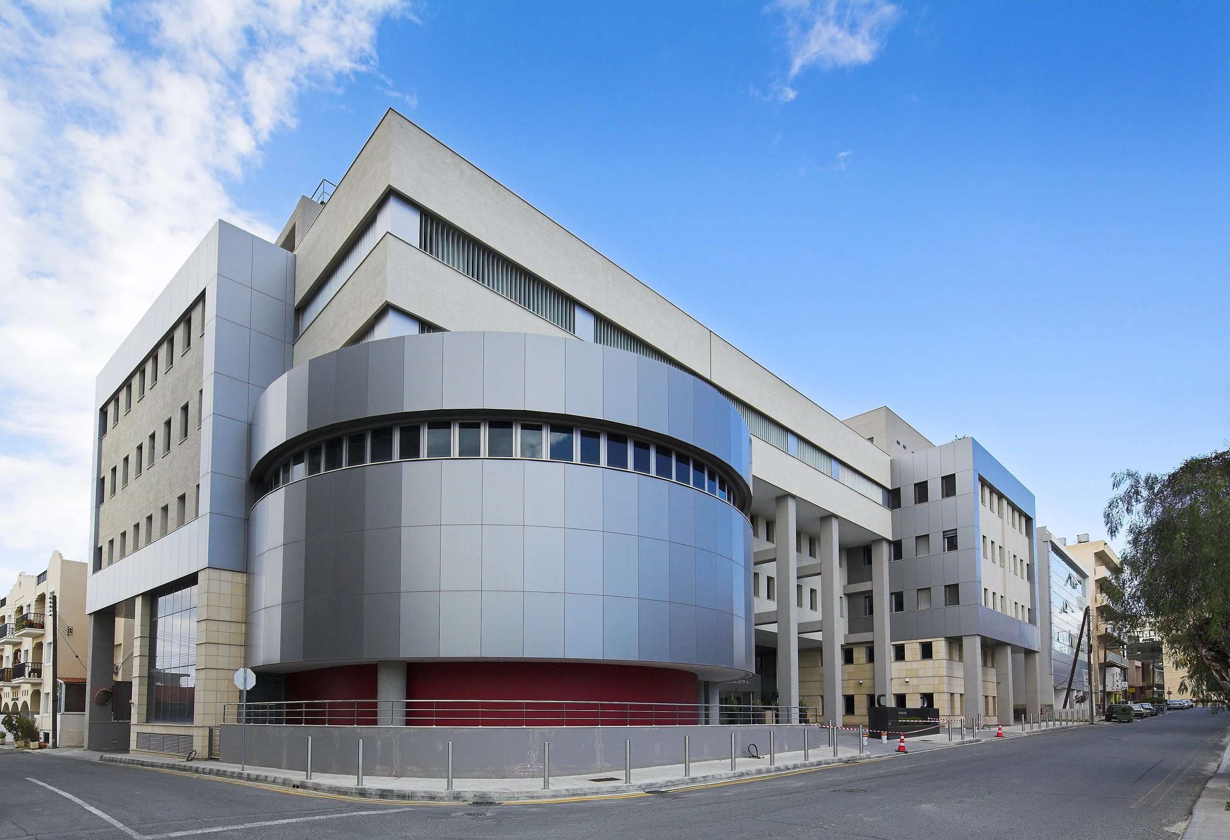 Modern multi-story building with curved and angular architectural design, large windows, and a clear blue sky with some clouds in the background.