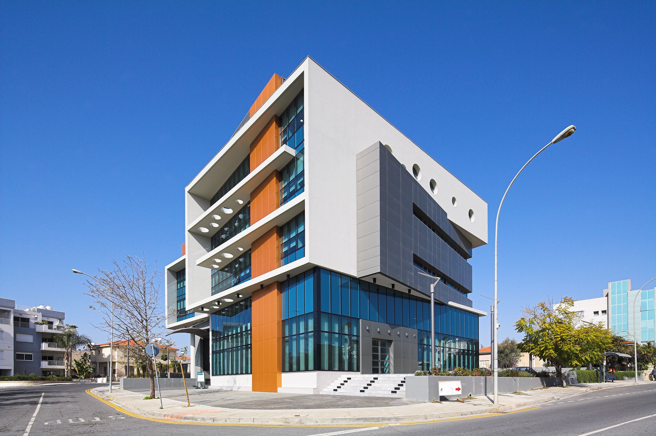 Modern multi-story building with glass windows and orange accents, located at a corner of a city street, under a clear blue sky.
