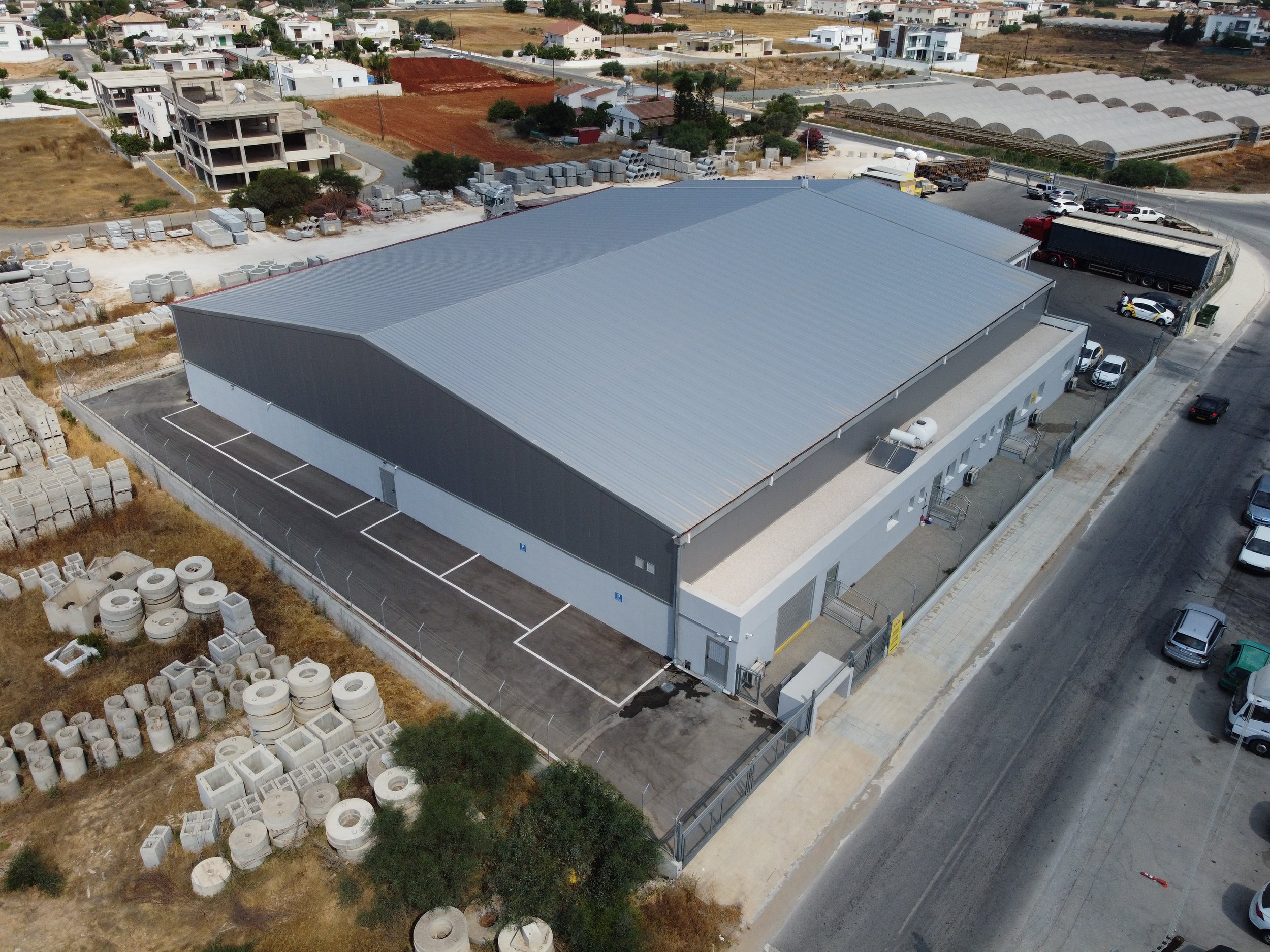 An aerial view of an industrial warehouse with construction materials, vehicles, and parked cars surrounding it, with residential buildings and greenhouses in the background.