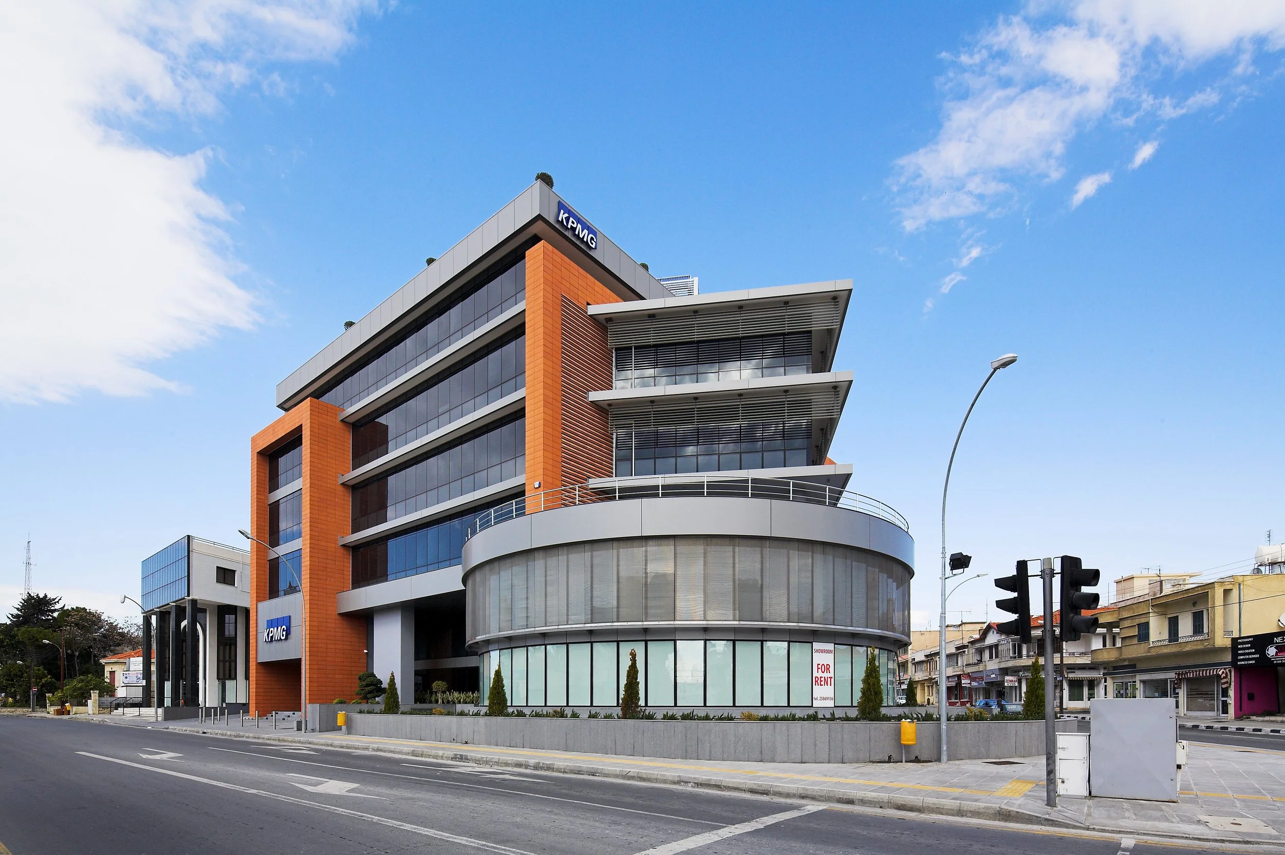 Modern multi-story building with glass windows and orange panels, with a sign for KPMG, located on a city street with traffic lights and sidewalks under a partly cloudy blue sky.