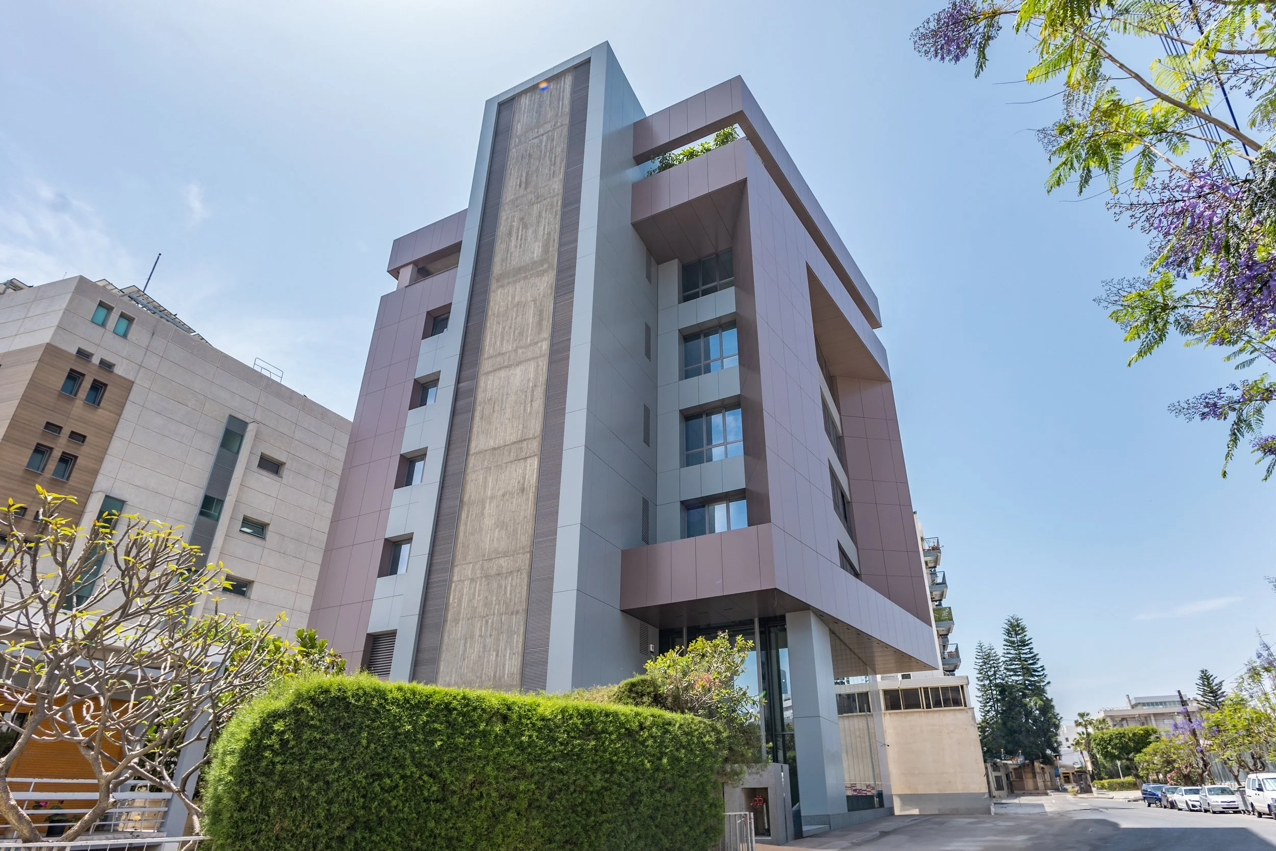 A modern multi-story building with a distinctive architectural design, featuring a blend of concrete and pastel-colored panels, with large windows and a landscaped area with shrubs and trees in the foreground.