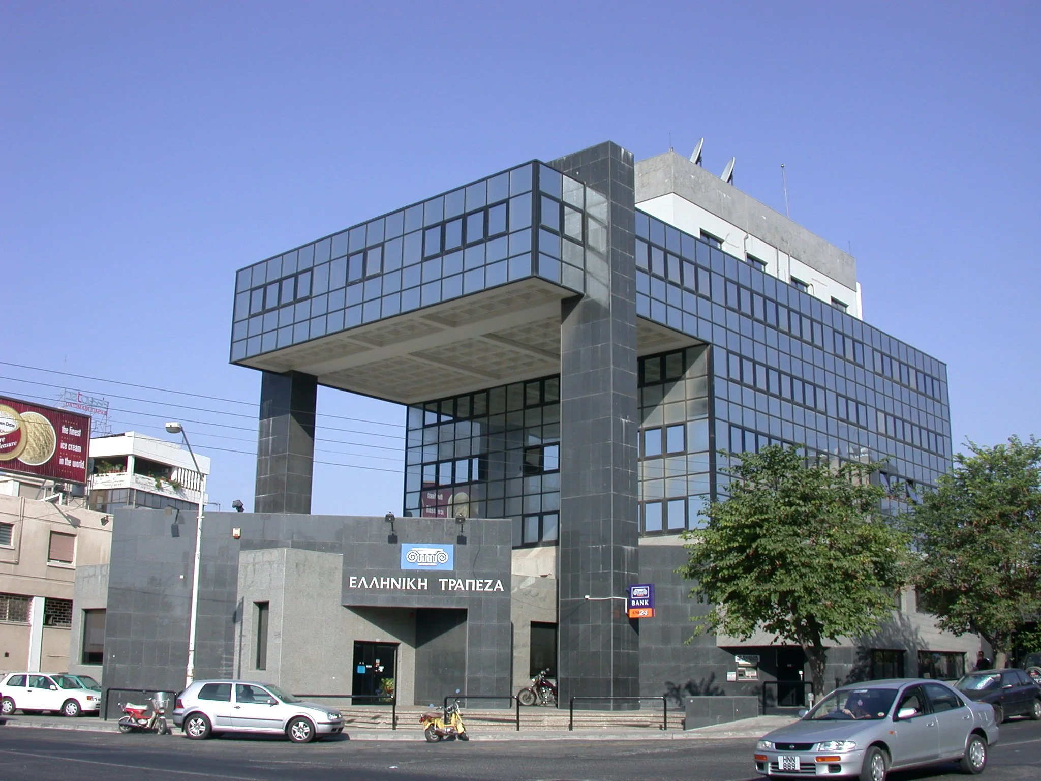 Modern gray office building with glass windows and Greek signage on the front, situated along a busy street with parked cars, motorcycles, and bicycles, under a clear blue sky.