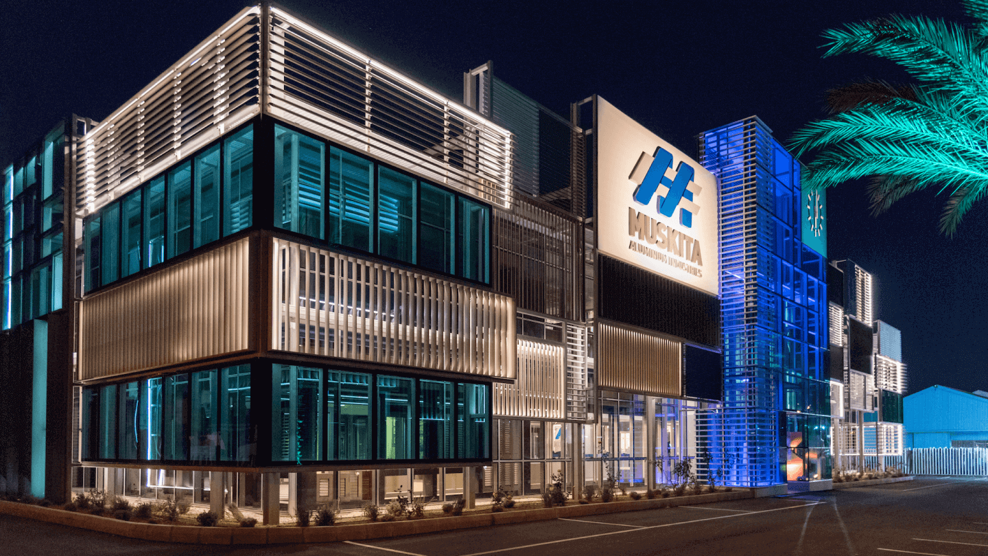 Night view of a modern building with illuminated windows, metal and glass exterior, and a large sign that reads "Muskita Aluminum Industries."