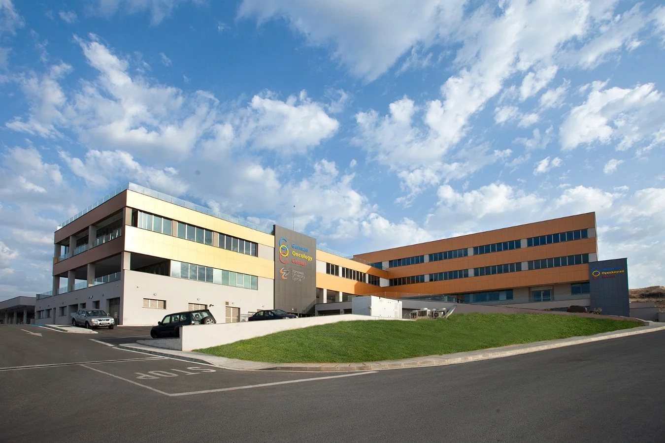 Modern multi-story healthcare building with colorful signage, parked cars, and a grassy slope under a partly cloudy sky.