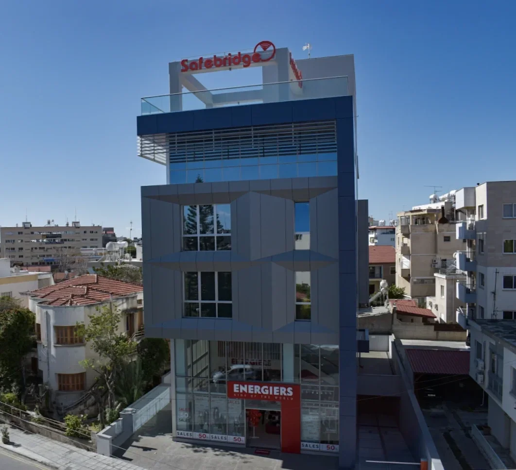 A multi-story modern building with a glass and metal facade situated among residential buildings. The top of the building features a red sign for Safebridge and a logo, while the ground floor has a sign for Energiers indicating a sales area.
