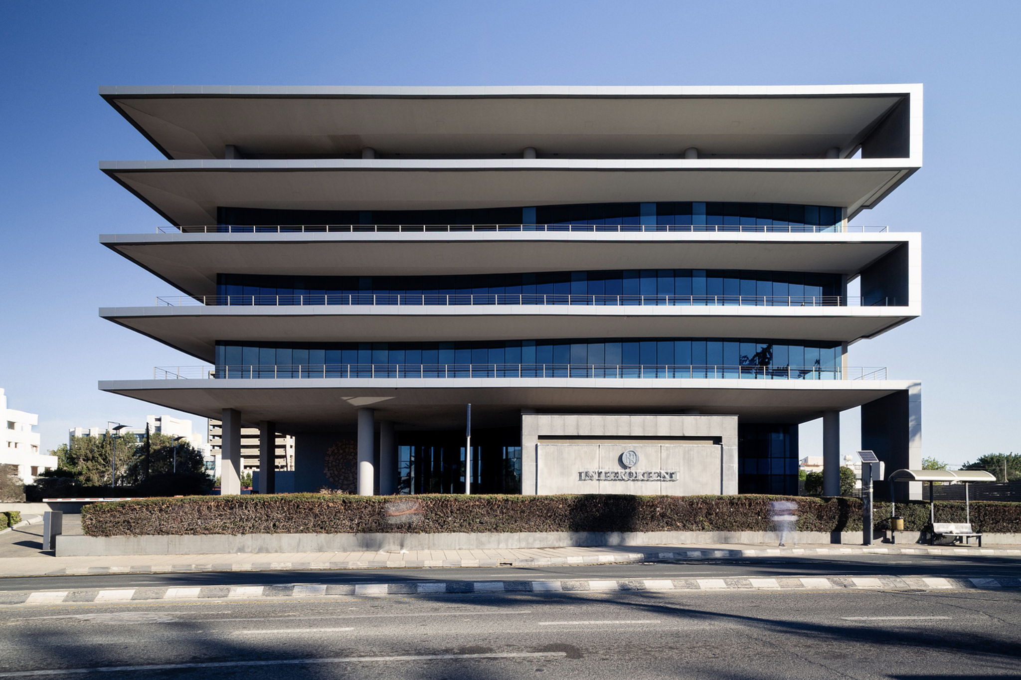 Modern multi-story building with overhanging white balconies, glass windows, and a concrete facade, with a landscaped foreground and a clear blue sky.