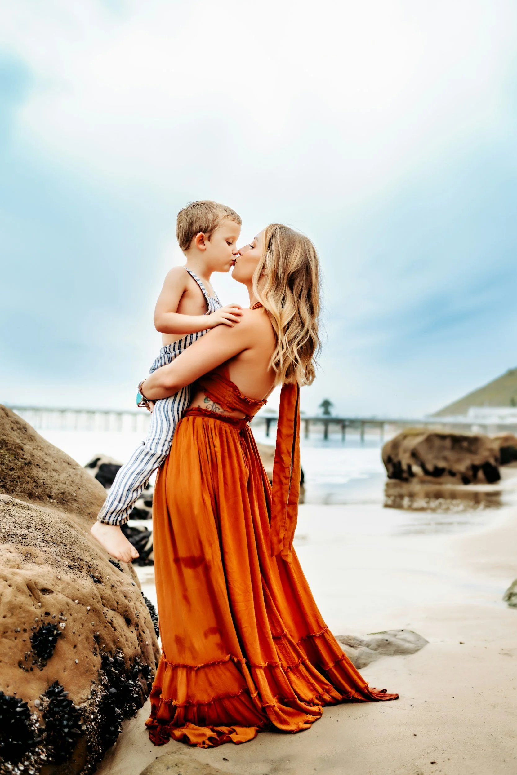 Una donna con un lungo abito arancione tiene un bambino in braccio sulla spiaggia, si baciano con lo sfondo del mare e del cielo nuvoloso.