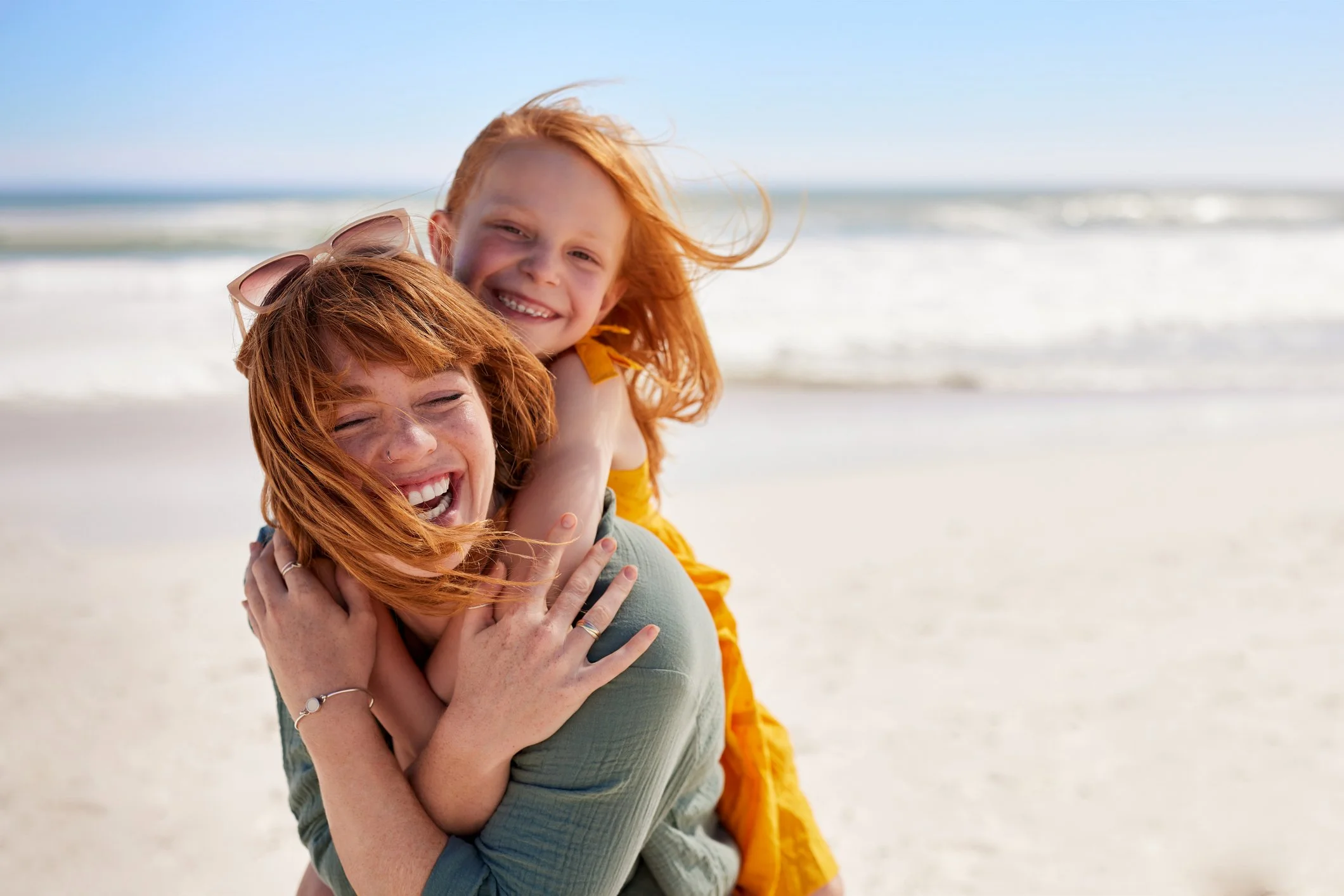 Due donne con i capelli rossi sorridono e si abbracciano sulla spiaggia con il mare e il cielo azzurro sullo sfondo.