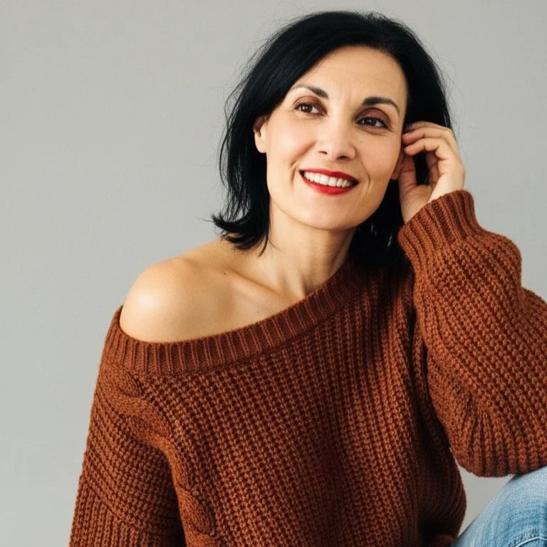 Ragazza con capelli neri, sorriso, maglione marrone a collo largo, posa con mano sulla testa su sfondo grigio.