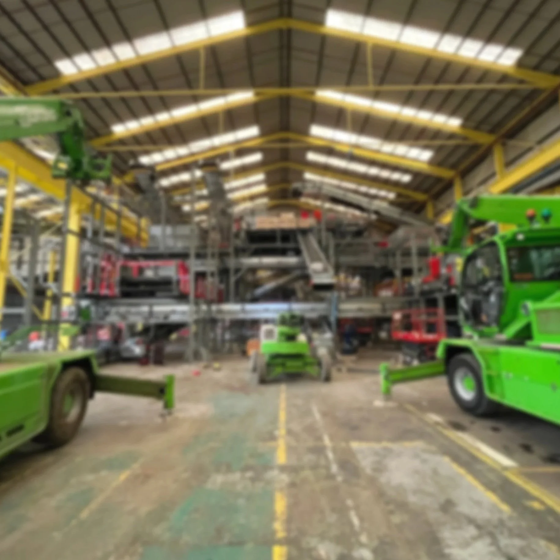 Indoor industrial warehouse with green heavy machinery, forklifts, and construction equipment parked inside.