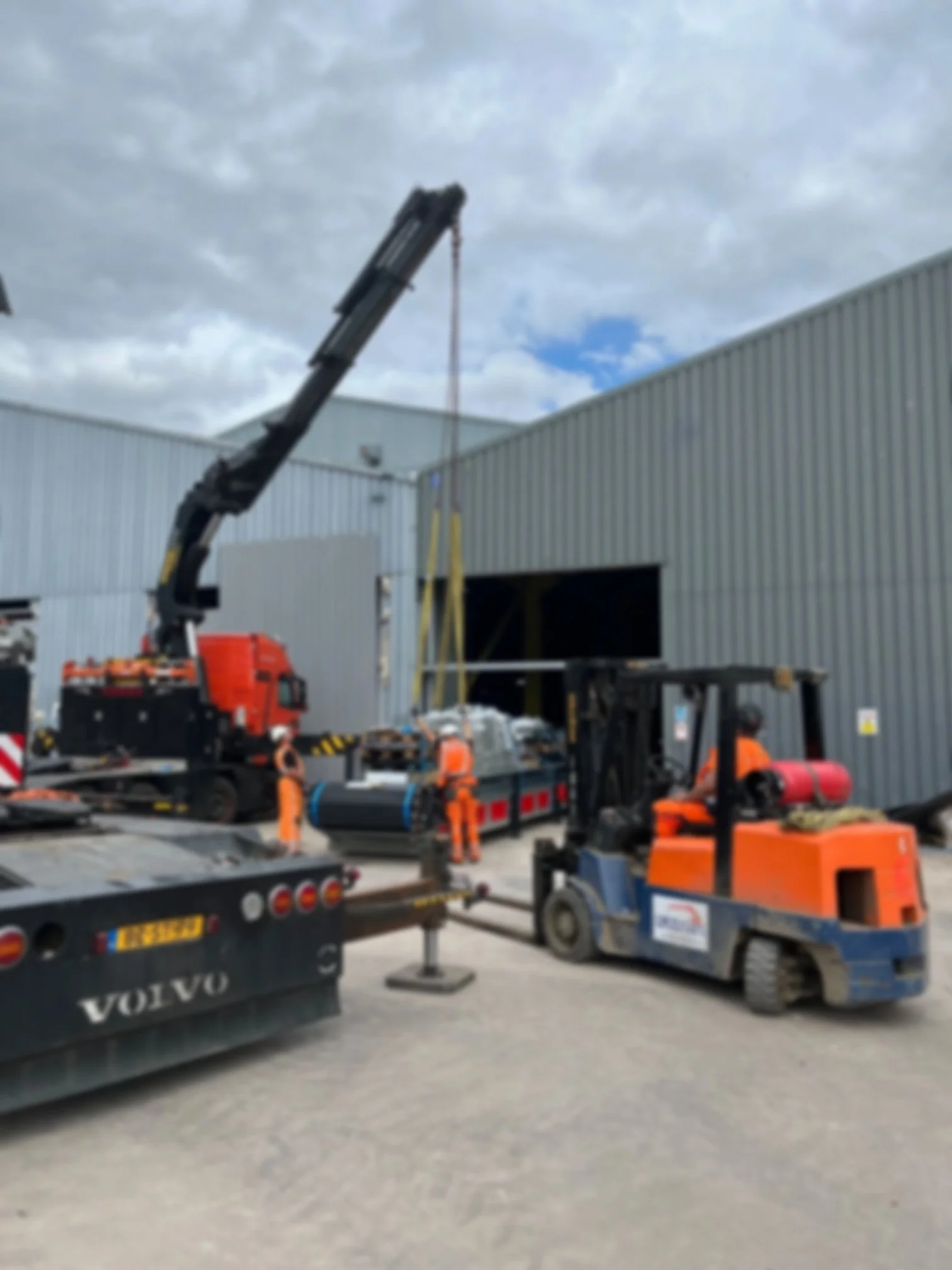 Construction workers using heavy machinery, including a crane and forklifts, to assemble or repair the exterior of a large industrial building.